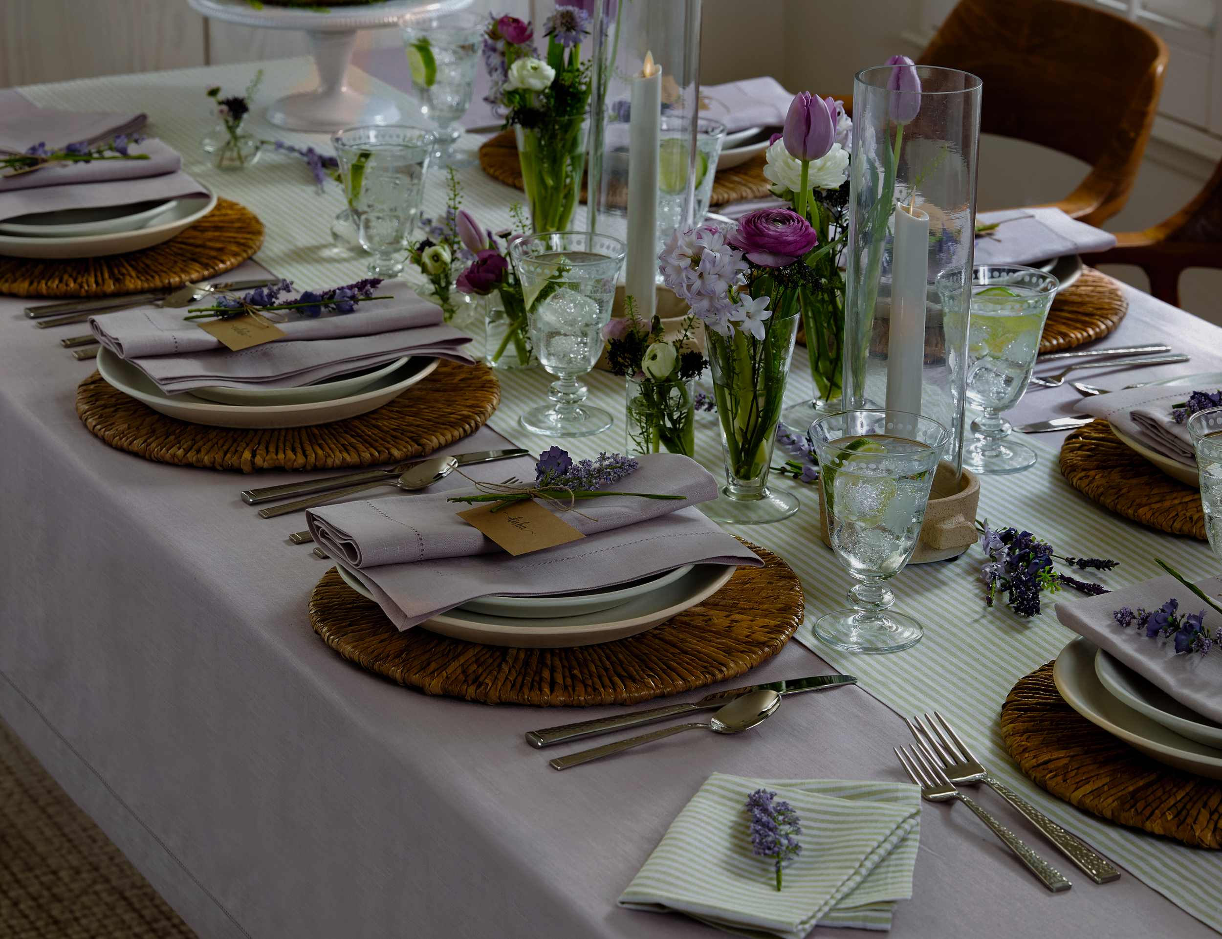 A beautifully set dining table with a white tablecloth, woven placemats, and lavender-themed decorations including fresh flowers in vases, candles, and lavender sprigs on napkins and tables.
