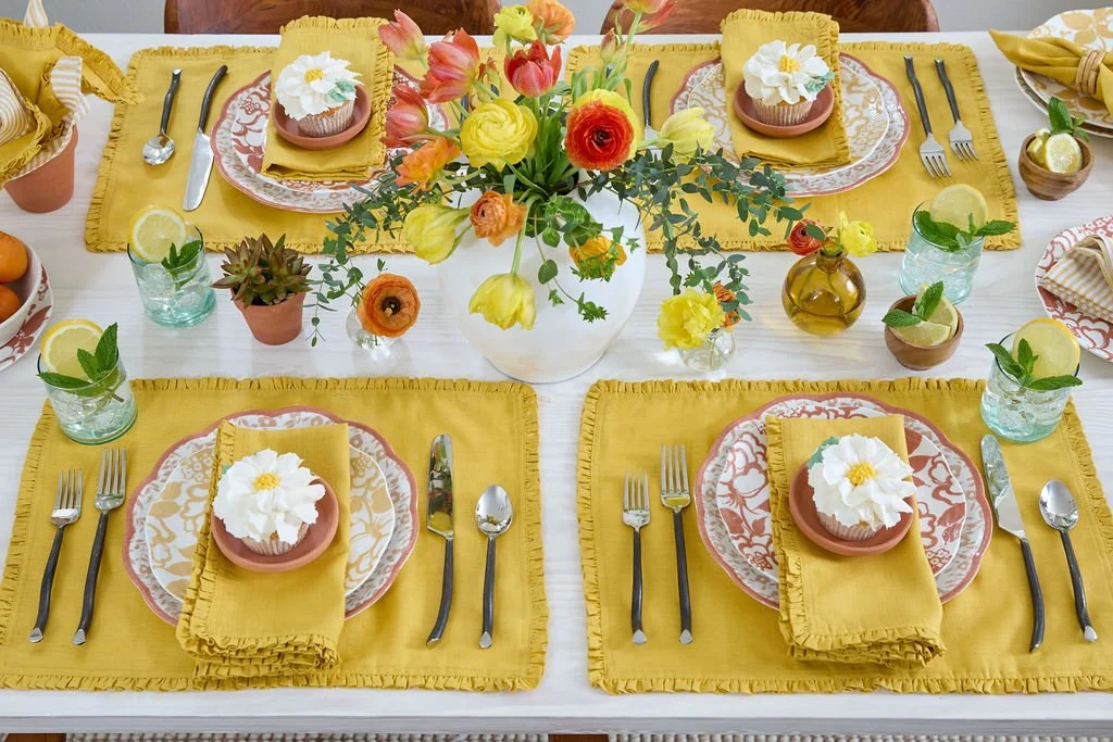 A table set for a meal with yellow placemats, floral patterned plates, yellow napkins, and cupcakes with white frosting and flower decorations. A centerpiece of mixed yellow, orange, and red flowers in a white vase is in the middle. There are glasses with lemon slices and mint leaves, small potted plants, and a small amber bottle on the table.