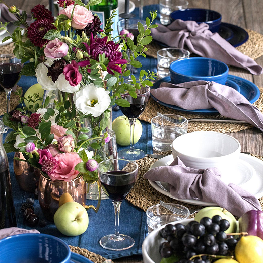 A table set with a large floral centerpiece, wine glasses filled with red wine, blue bowls, white plates, and cloth napkins, with apples and grapes as snacks.