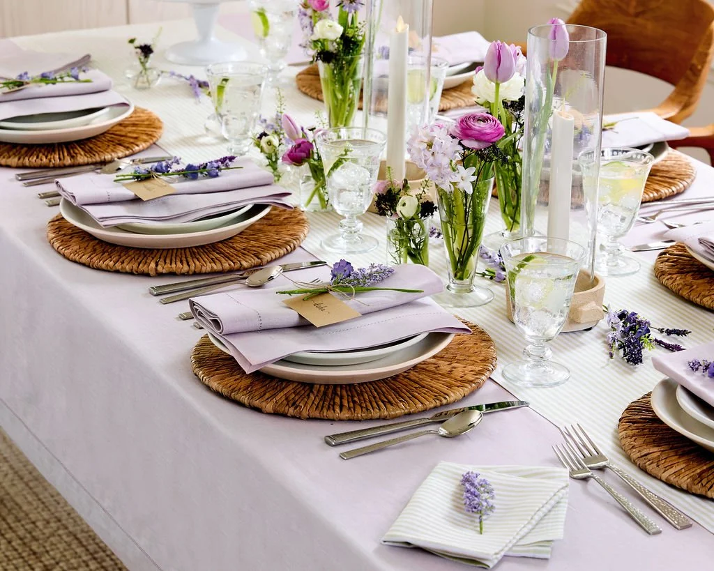 Elegant dining table set with white tablecloth, wicker placemats, white plates, lavender napkins, glassware, and centerpieces of pink and purple flowers with white candles.