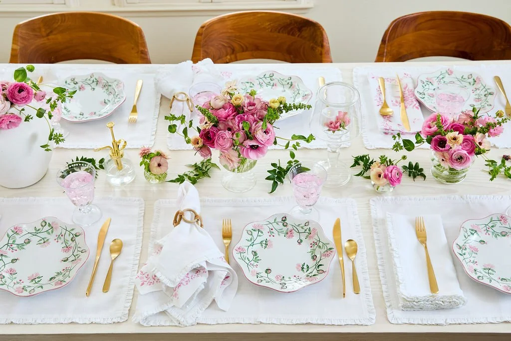 A dining table decorated with pink and white flowers, gold flatware, floral-patterned plates, and pink-tinted glasses, set for a celebration.
