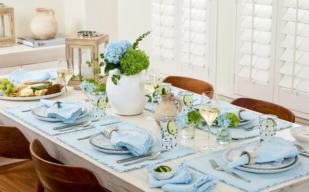 A dining table decorated for a meal with a blue and white theme, featuring plates, glasses, napkins, and a large white vase filled with blue and green flowers.