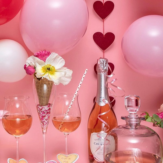 Decorative setup with pink balloons, a bottle of rosé wine, two glasses of rosé, flowers in a cone-shaped vase, and hanging red paper hearts against a pink background.