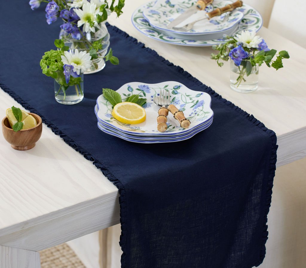 A dining table with a navy table runner and white tablecloth, decorated with floral arrangements in small vases, a stack of decorated plates, lemon slices, fresh mint leaves, and bamboo-handled cutlery.