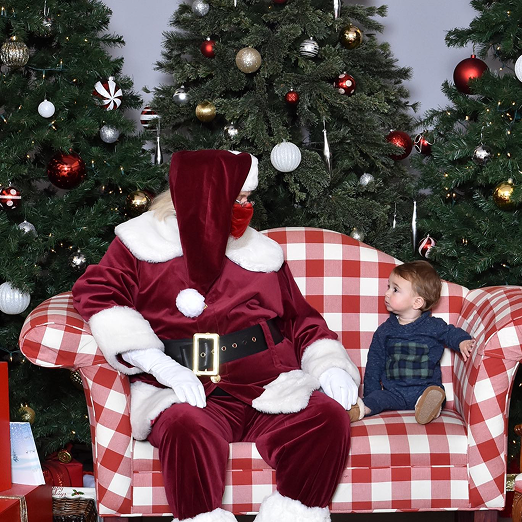 A young child sitting on a red and white checkered couch with Santa Claus, who is dressed in a traditional red suit with white fur trim, in front of decorated Christmas trees with ornaments and lights.