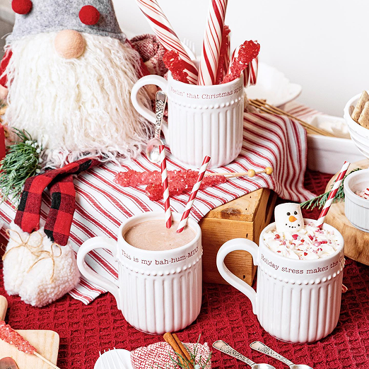 Three white ceramic mugs with holiday-themed text, filled with hot chocolate and candy canes, surrounded by Christmas decorations on a red textured tablecloth.