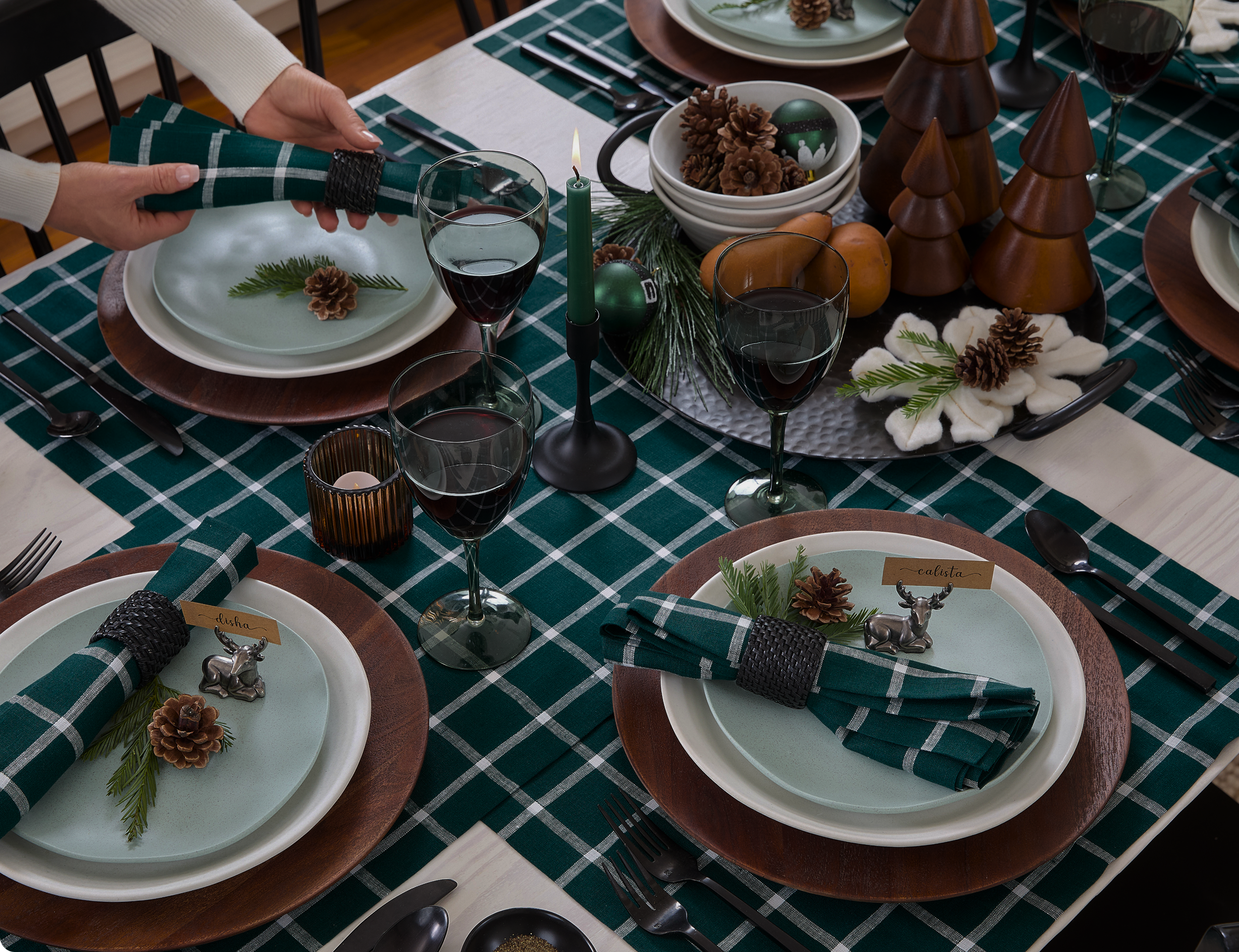 A festive Christmas table setting with green checkered tablecloth, decorated with pinecones, sprigs of pine, candles, and holiday ornaments. Each place setting includes a plate, a napkin with a pinecone and sprig, and a small reindeer decoration during preparation.