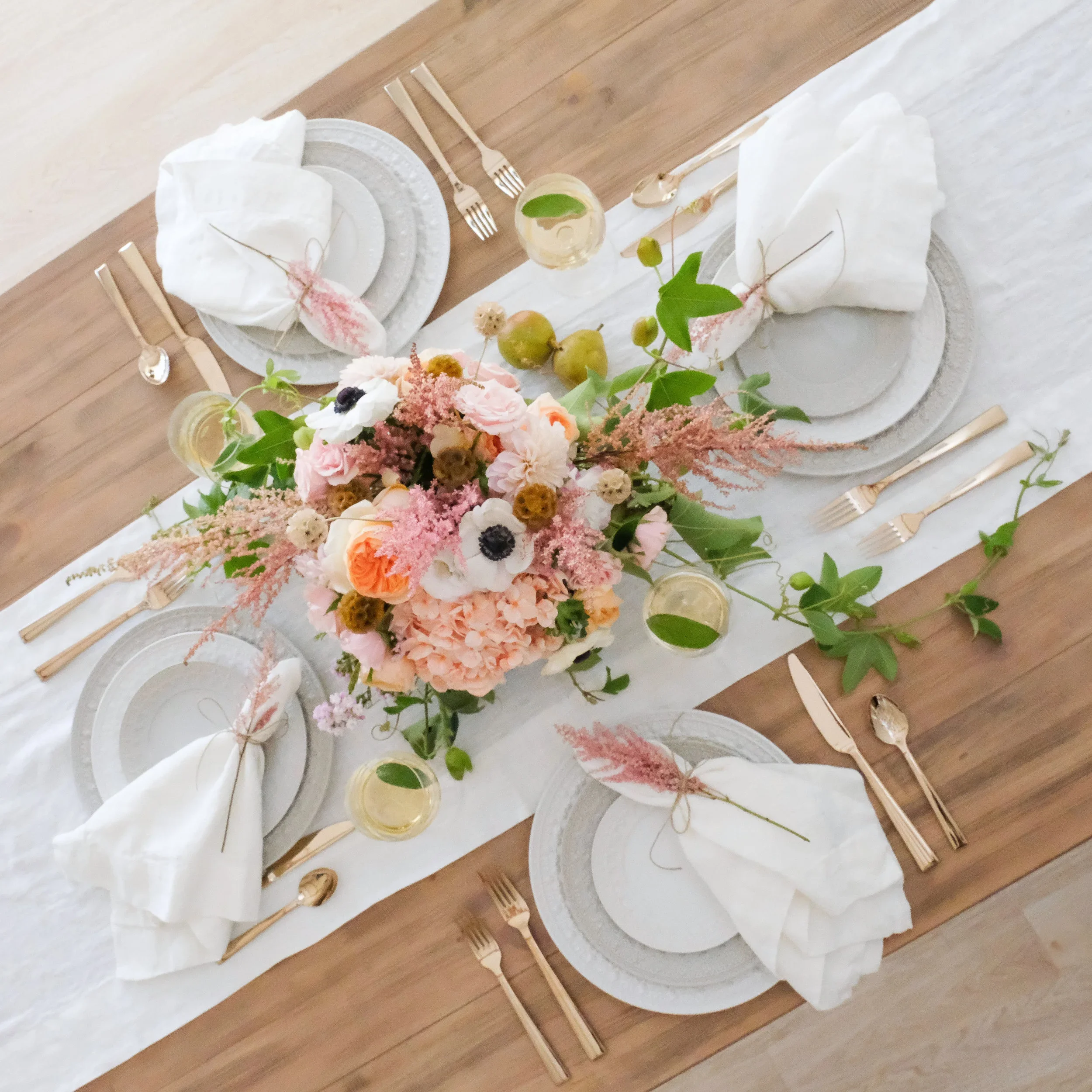 A table set for a formal dinner with a floral centerpiece, white napkins, gold utensils, clear glasses with drinks, and a white table runner.