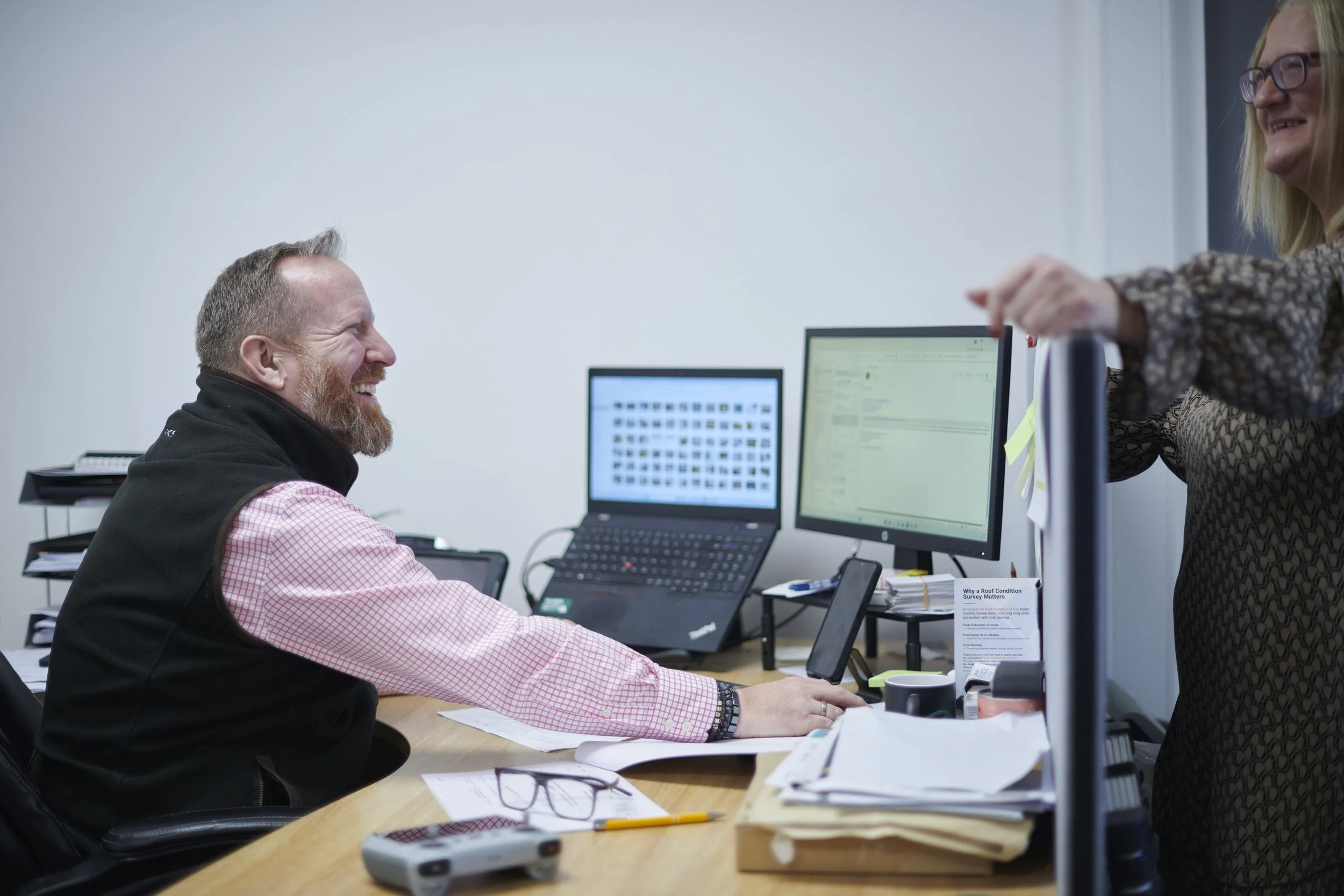 Two colleagues laughing and chatting in an office, one sitting at a desk with multiple monitors, the other standing next to her.