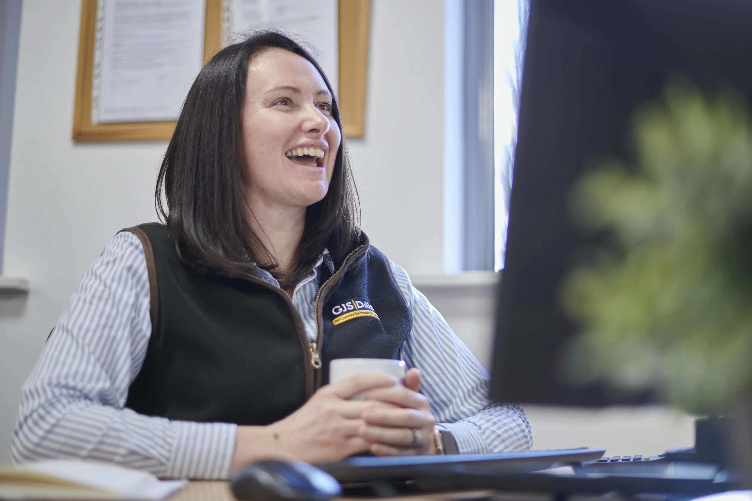 A woman with dark hair, wearing a GJS vest over a striped shirt, sitting at a desk with a computer, smiling and holding a white mug in an office setting.