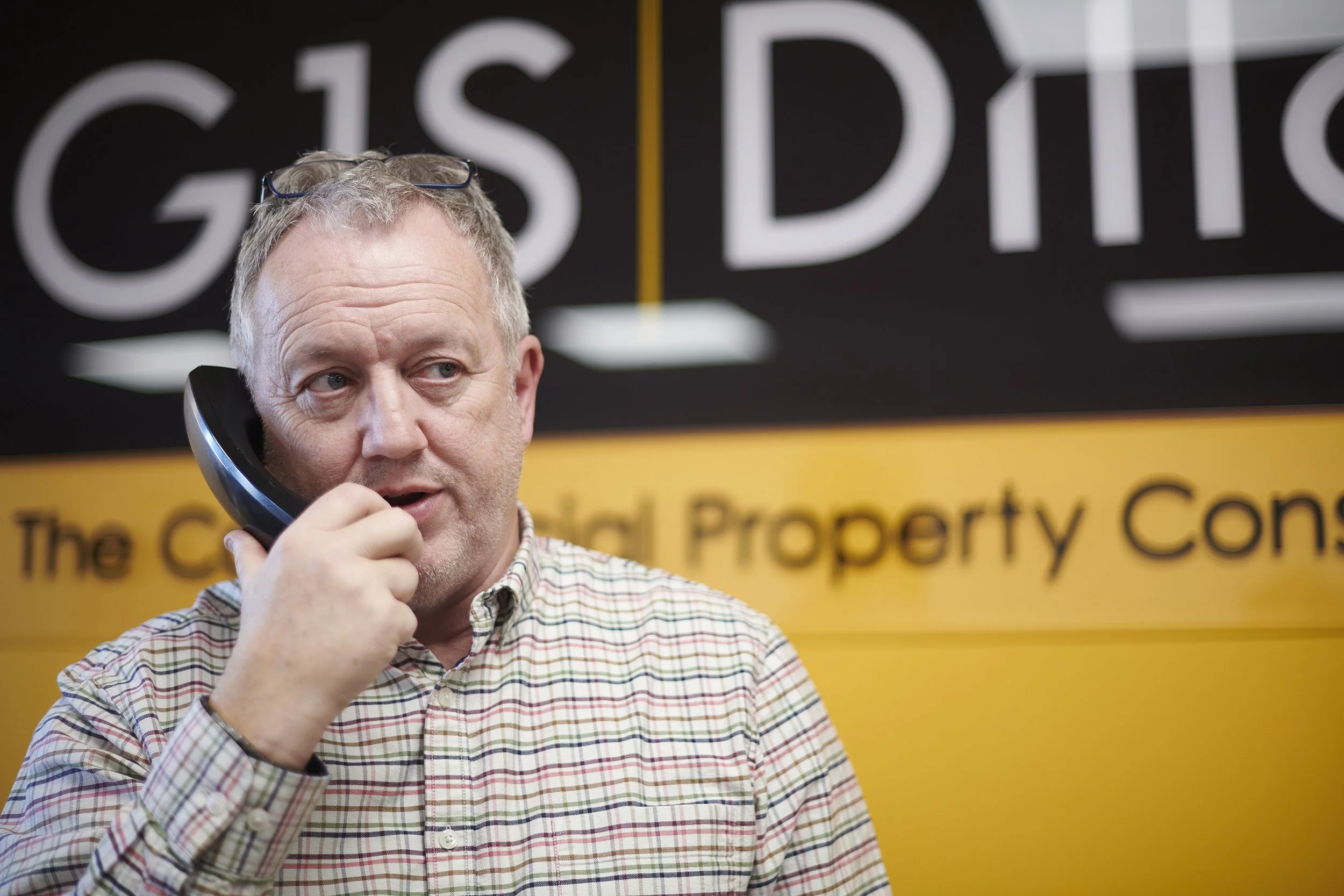 A man with glasses and gray hair sitting at a desk in an office, holding a yellow pen, with papers and brochures on the table, having a conversation.