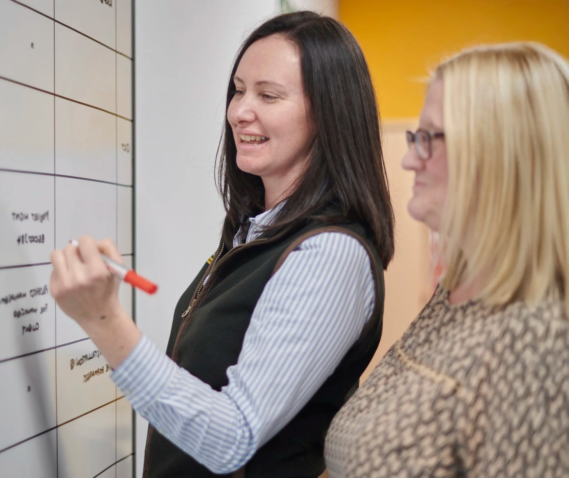 Two women standing at a whiteboard with writing, one writing with a red marker, smiling, and the other observing, in a professional setting.