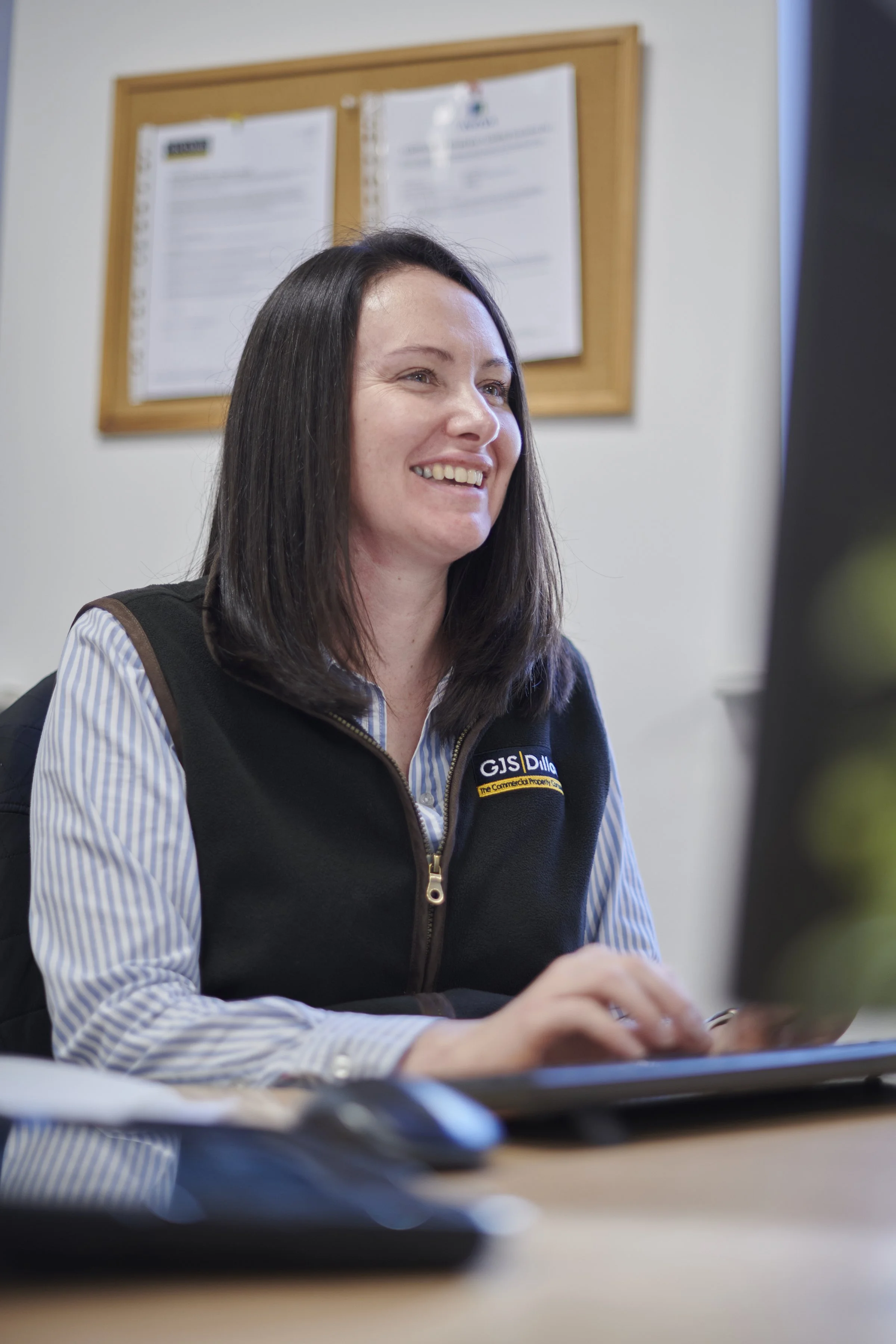 A woman with dark hair smiling and working at a computer in an office setting.