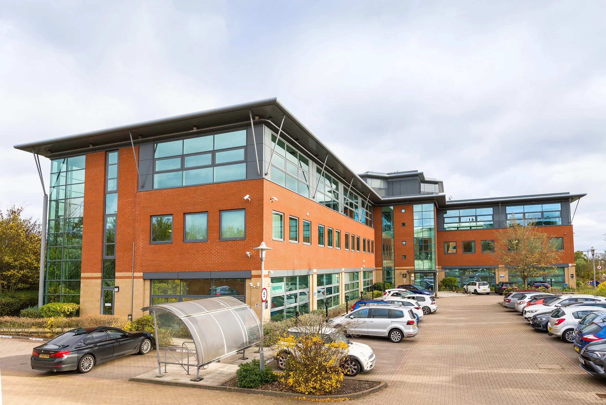 Modern office building with large glass windows, brick and metal exterior, parking lot with cars, and a bike shelter in front.