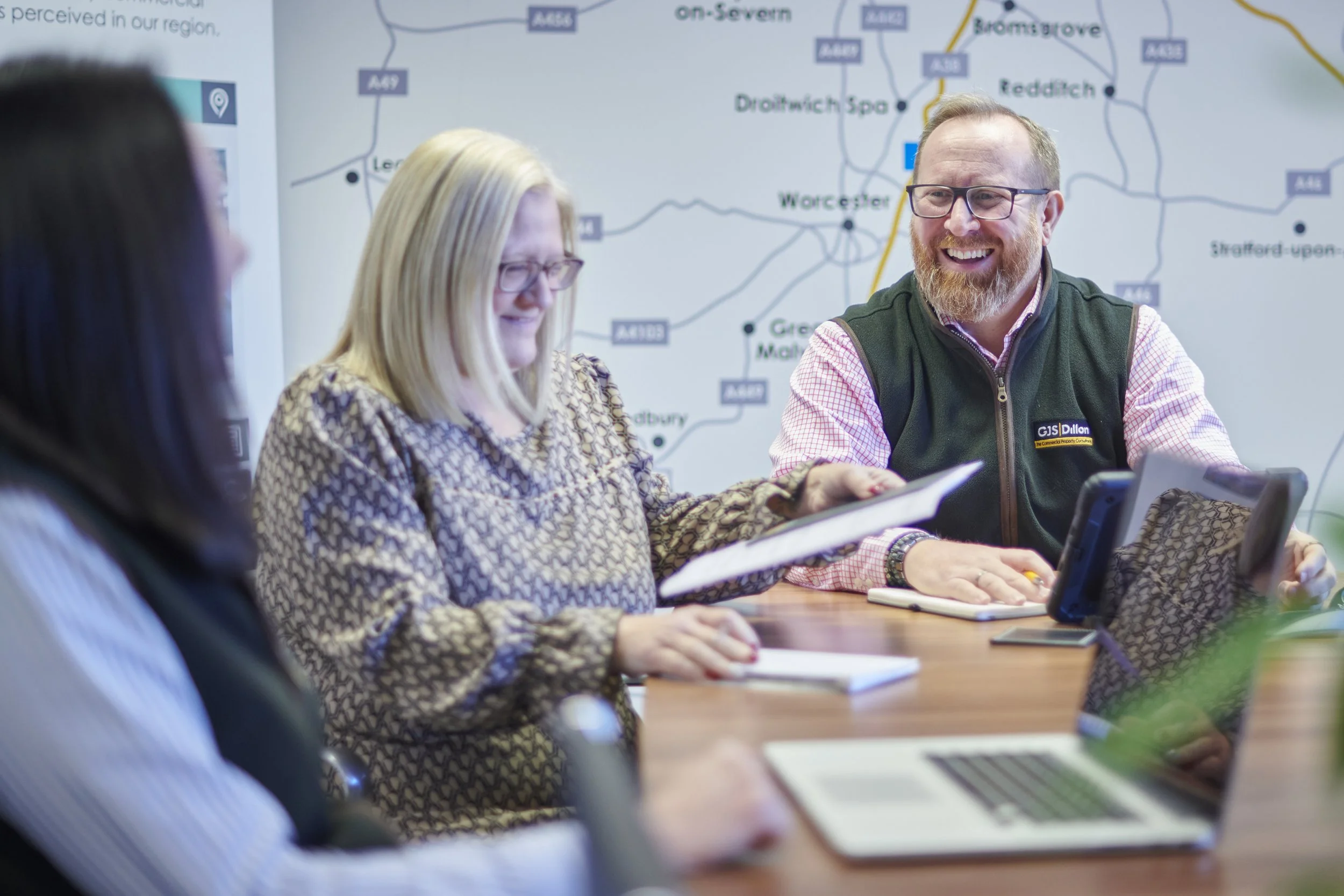 Three people sitting at a table in a meeting room, smiling and talking, with a large map of the Worcester area in the background.