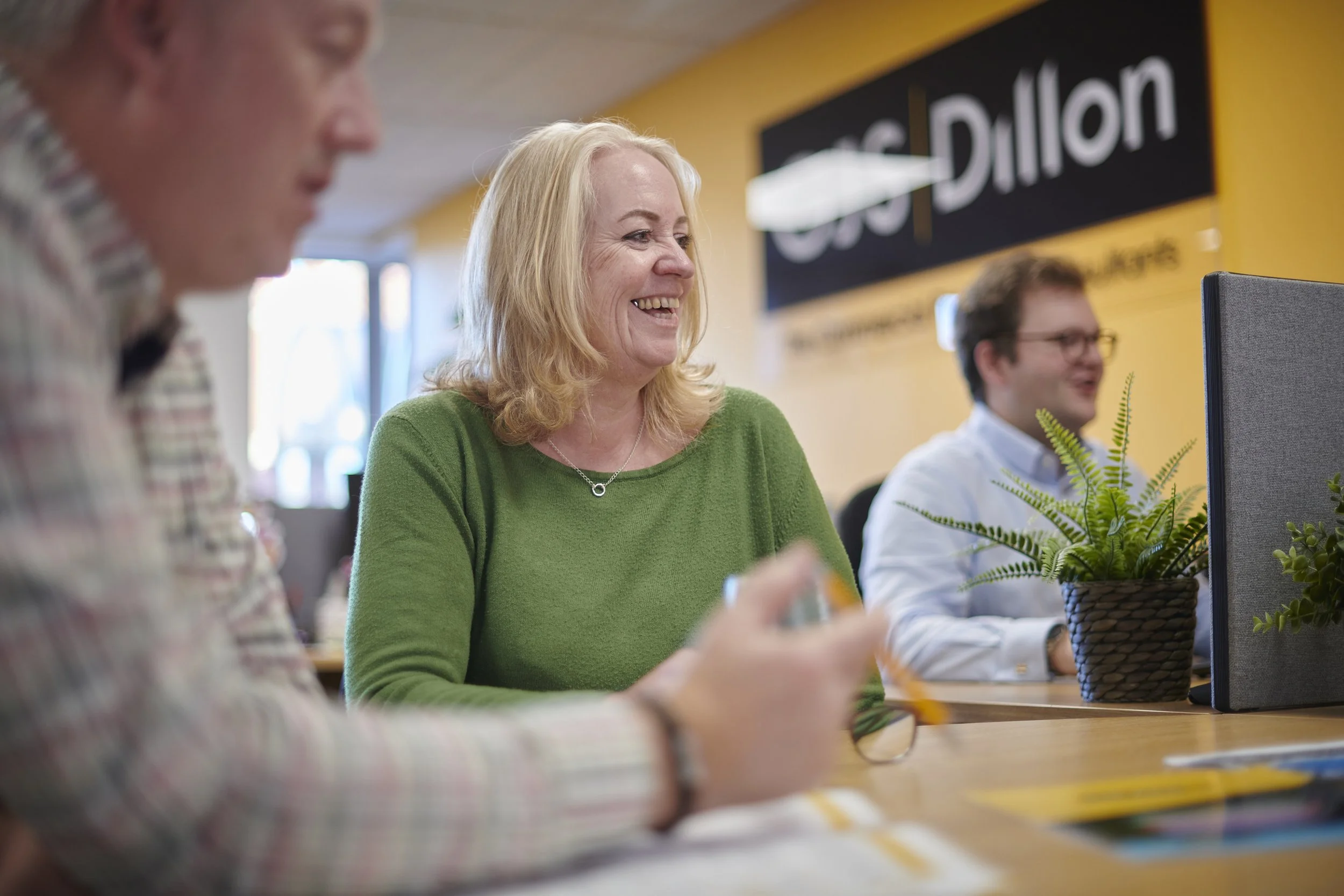 People sitting at a desk in an office, smiling and working, with plants on the desk and a sign reading 'Crest Dillon' in the background.