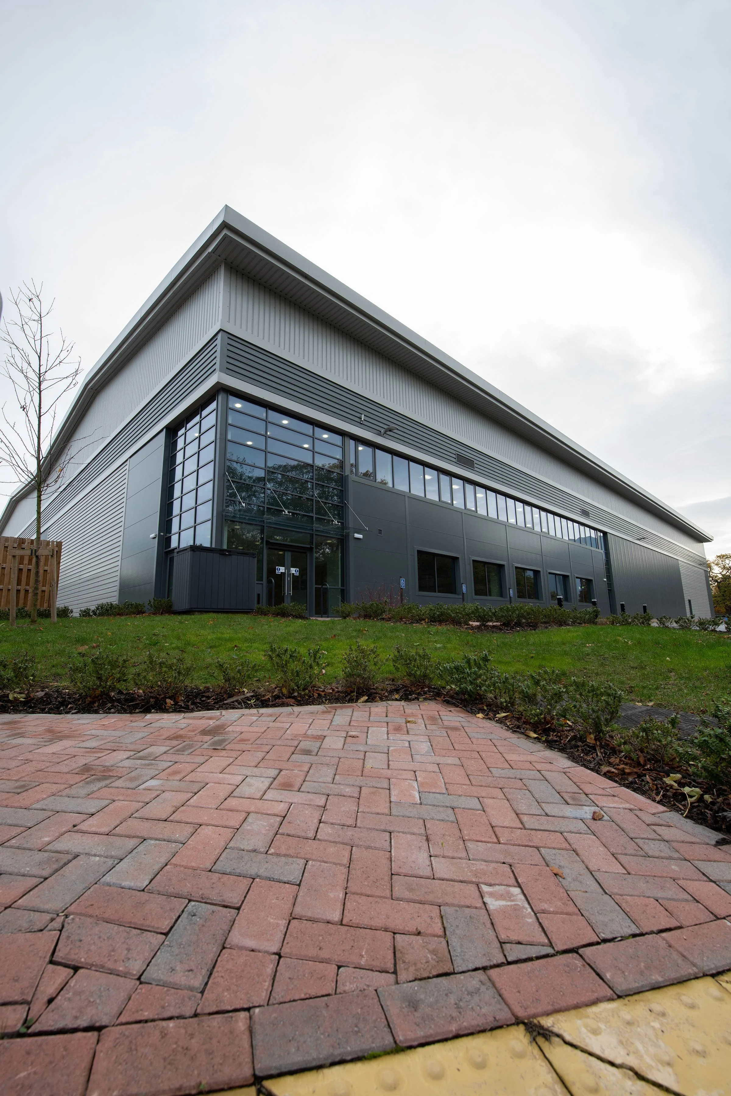 Modern multi-story building with large glass windows and metallic siding, surrounded by a small landscaped area with grass and shrubs, on a cloudy day.