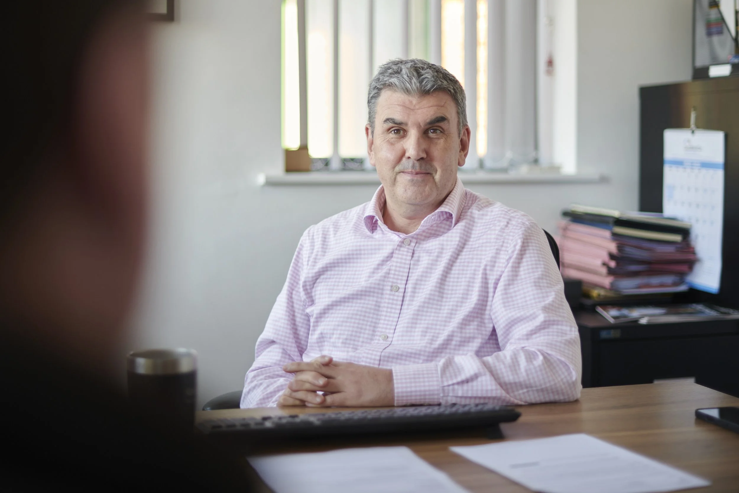 A middle-aged man with gray hair sits at a desk in an office, wearing a light pink checkered shirt. There are windows behind him and stacks of files on a shelf. His hands are clasped on the desk, and he looks at someone out of the frame.