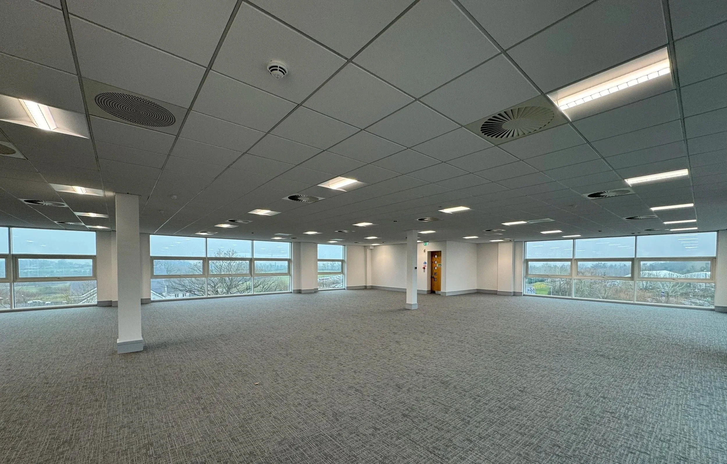 Empty office space with large windows, carpeted floor, and ceiling lights and vents.