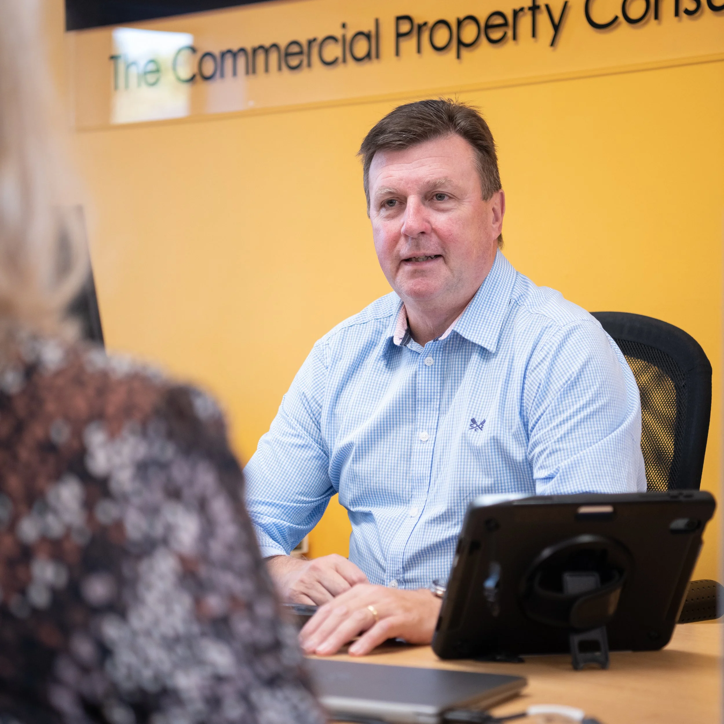 A man with short brown hair in a light blue checkered shirt sitting at a desk with a woman in the foreground. The man is in an office environment with a yellow wall and a sign that reads 'The Commercial Property Co.' in the background.