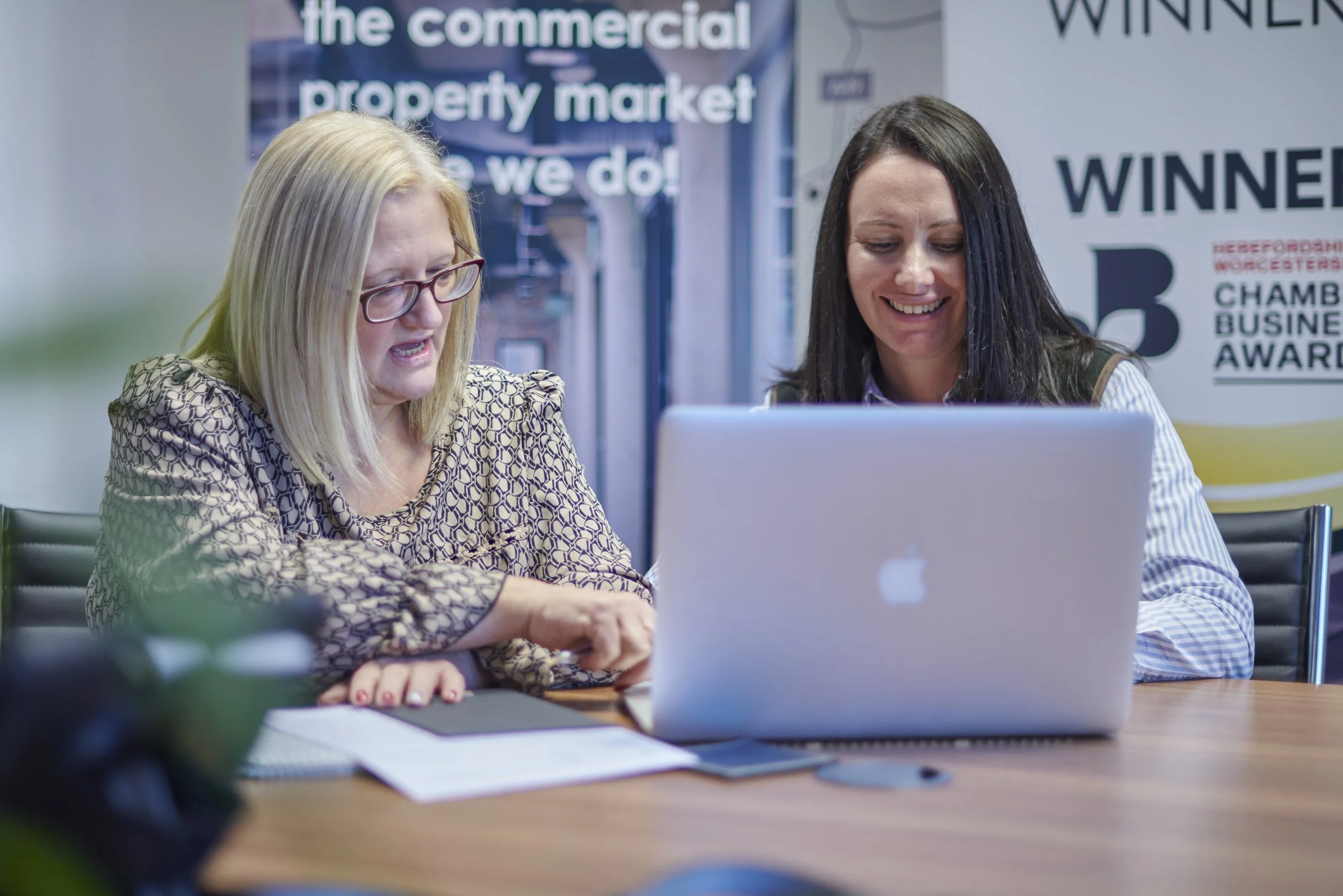Two women sitting at a conference table, looking at a laptop, with a banner behind them that mentions a business awards event.