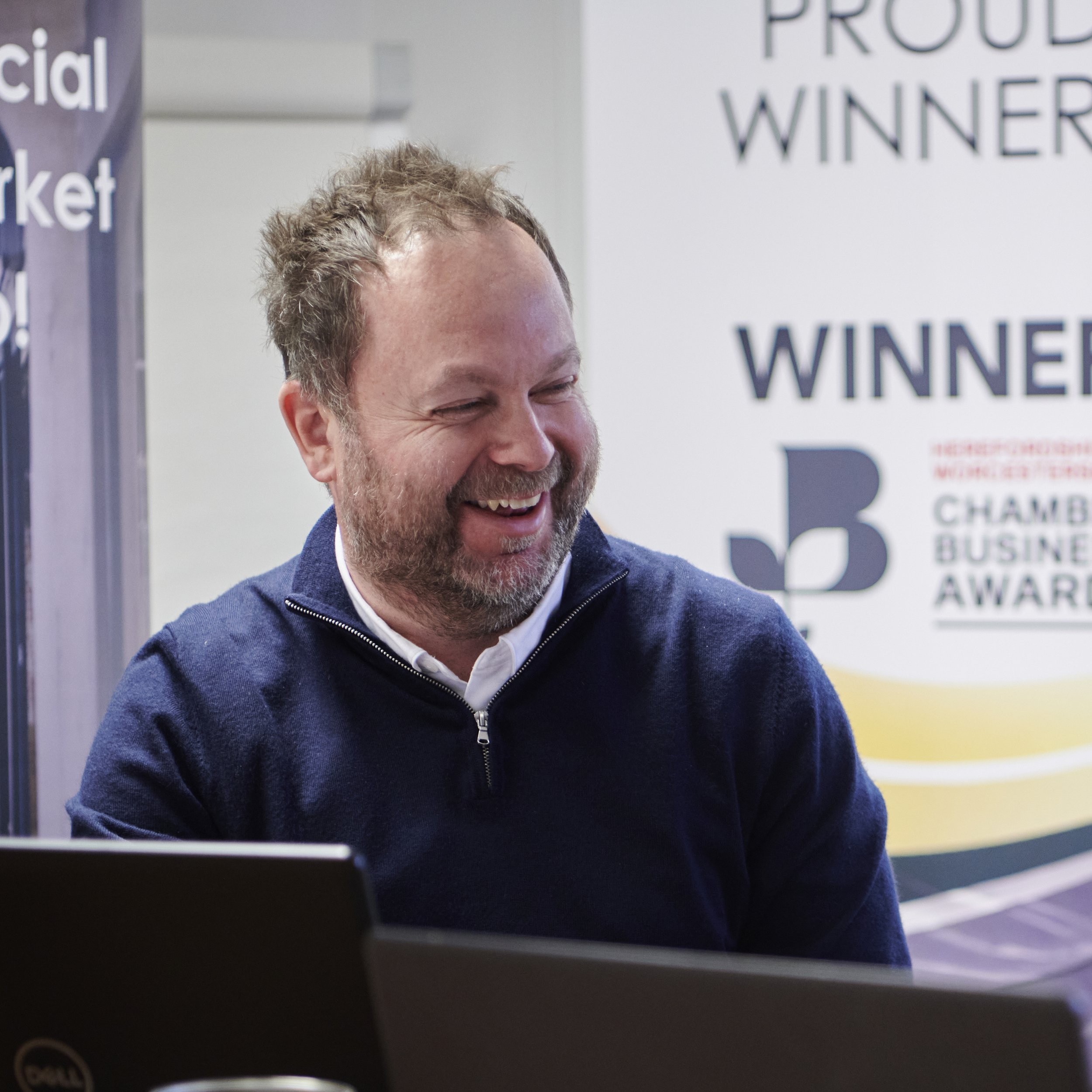 A man with a beard and short hair standing with arms crossed, wearing a light blue button-up shirt and dark pants, smiling against a plain white wall.