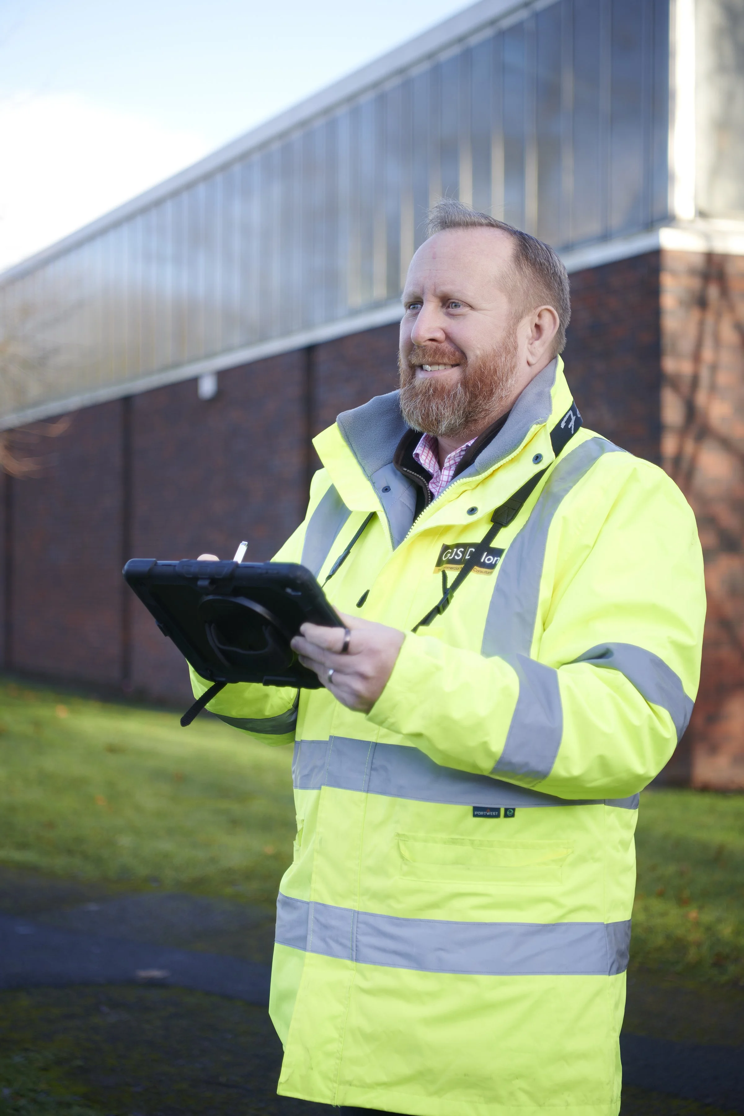 A man with a beard wearing a neon yellow high-visibility jacket and holding a tablet outdoors.