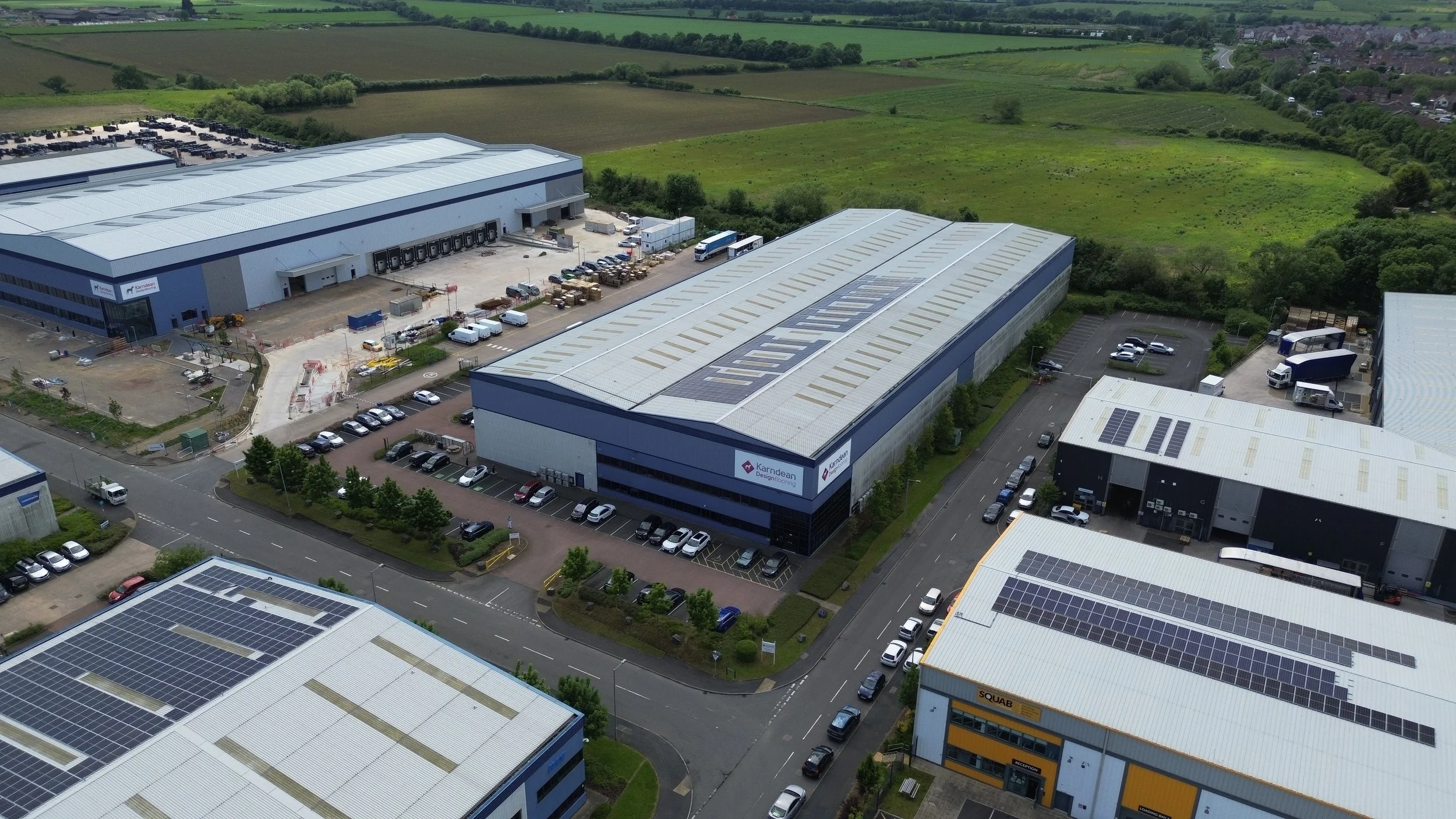 Aerial view of a commercial industrial complex with multiple warehouse buildings featuring solar panel roofs, parking lots filled with cars, and surrounding green fields.