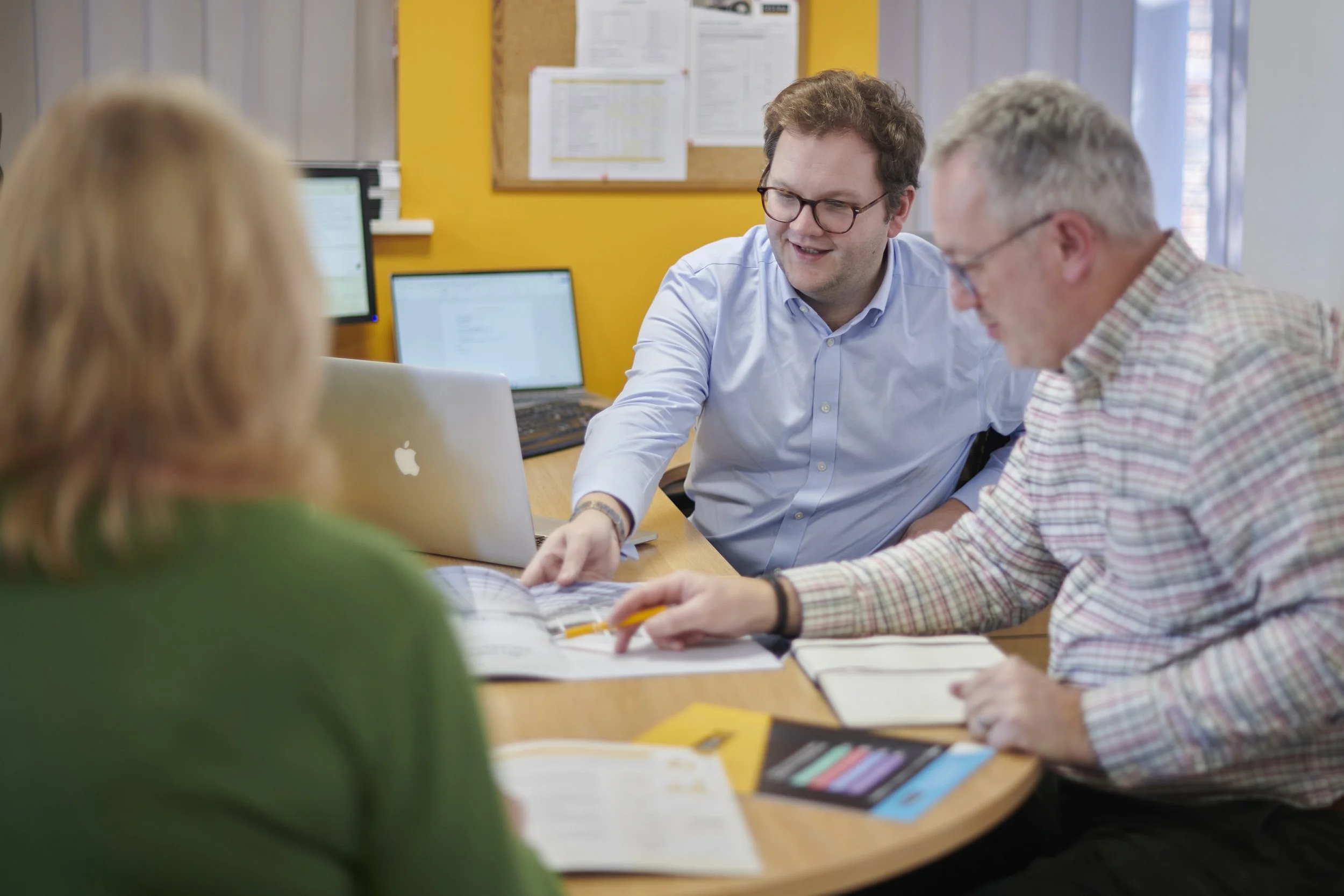 Three individuals engaged in a discussion at a meeting table with notebooks, a yellow brochure, and a laptop, in a room with computers and a yellow wall.