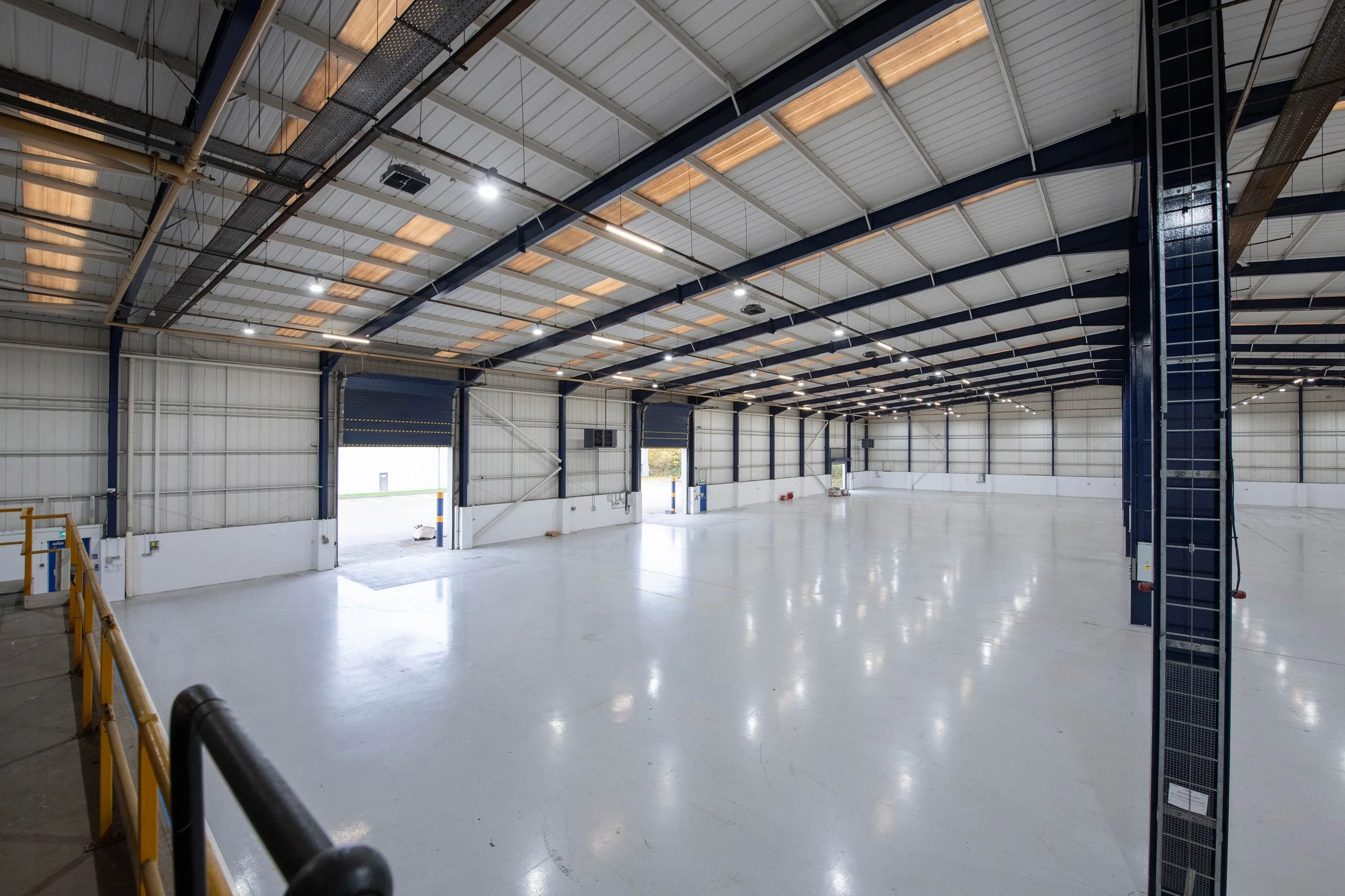 Empty indoor warehouse with high ceiling, metal beams, overhead lights, large open floor, and open garage doors.