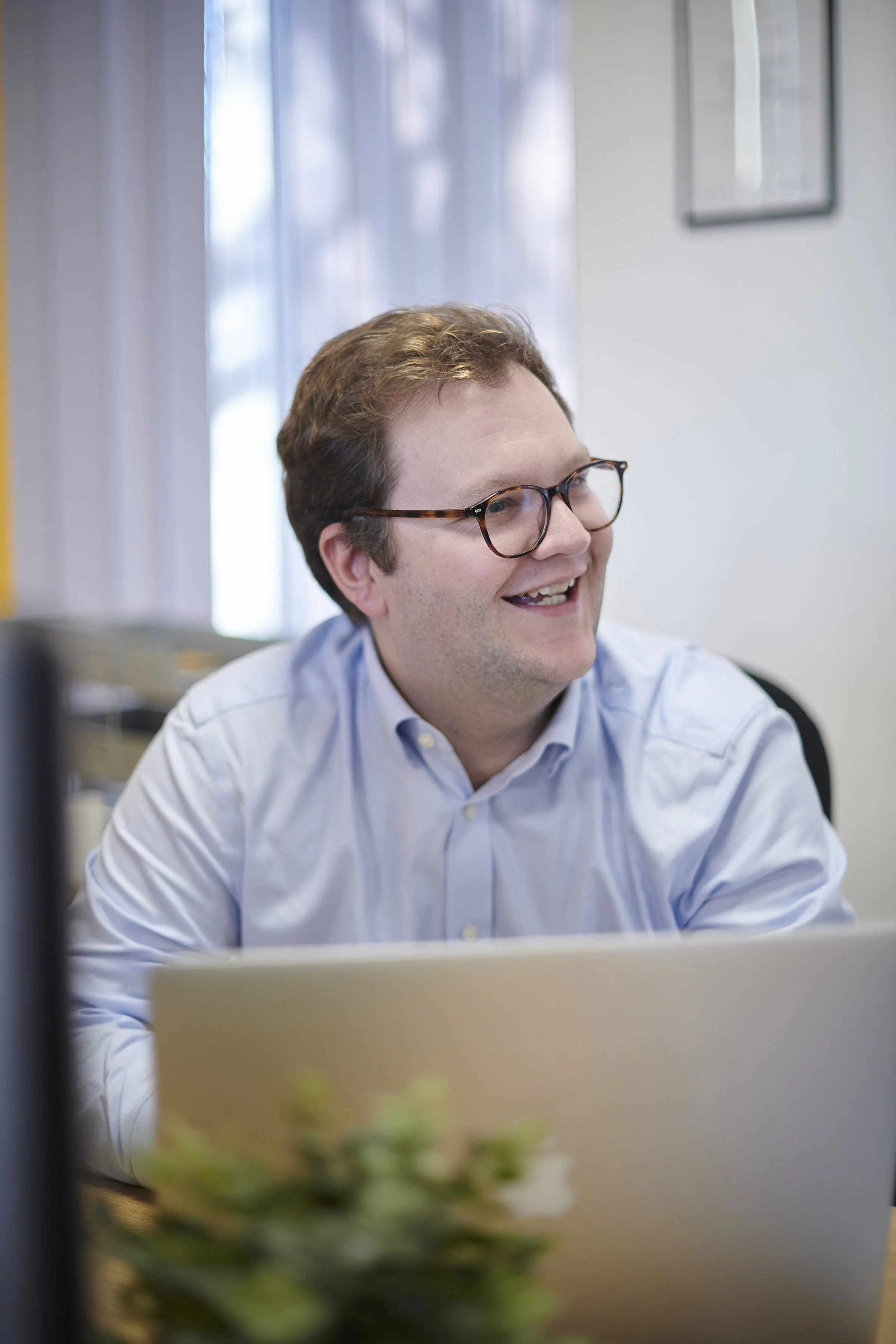 Man with glasses, smiling, working on a laptop in an office near a window.