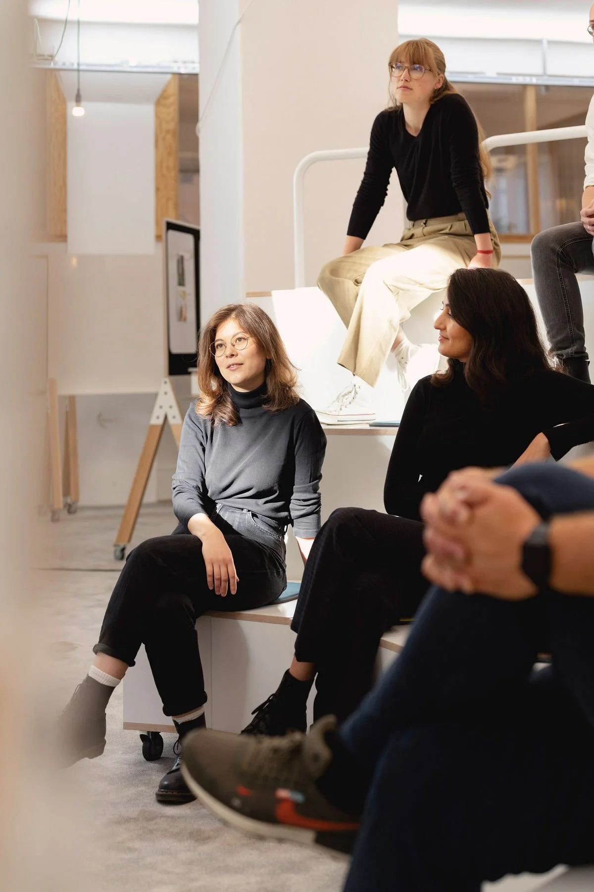 Un groupe de femmes assises et écoutant lors d'une réunion ou d'une discussion en intérieur modern, avec un fond de mur blanc et des présentoirs.