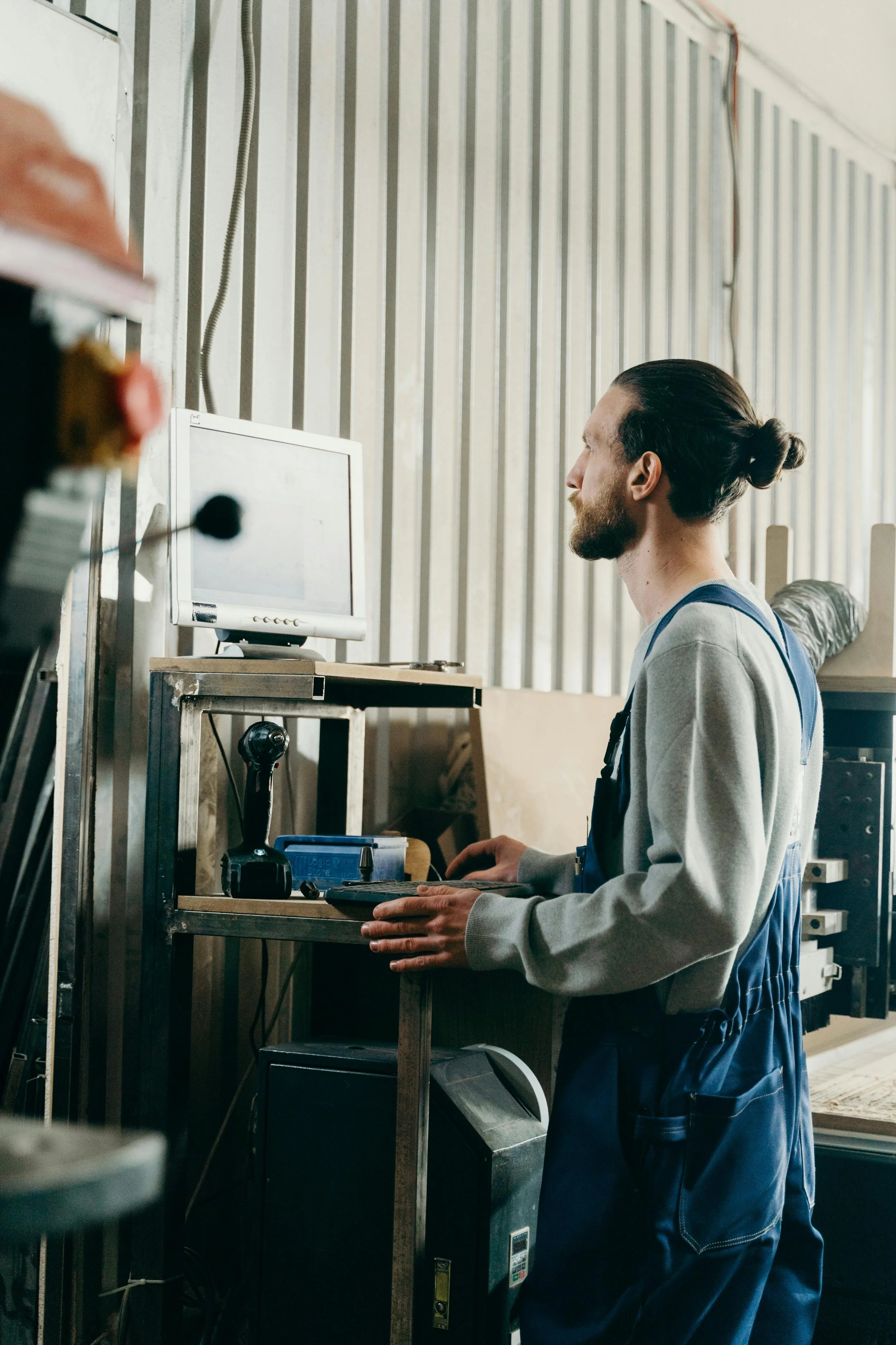 Un homme avec des cheveux longs attachés en chignon, portant une chemise grise et un pantalon de travail bleu, travaille dans un atelier avec un ordinateur sur un bureau en métal et plusieurs outils autour.