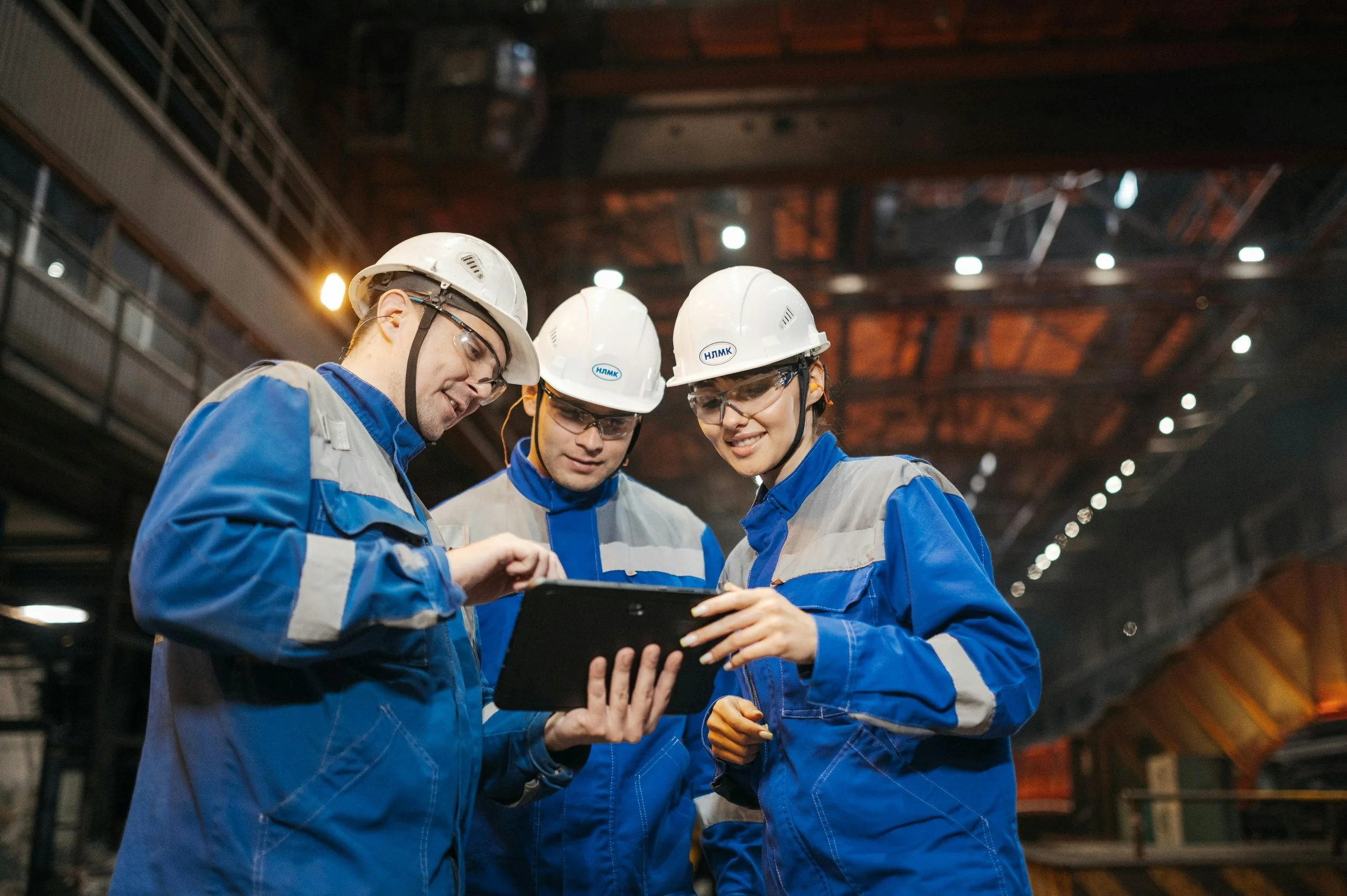 Trois travailleurs en uniformes bleus et casques de sécurité regardant une tablette dans une usine ou un atelier industriel.