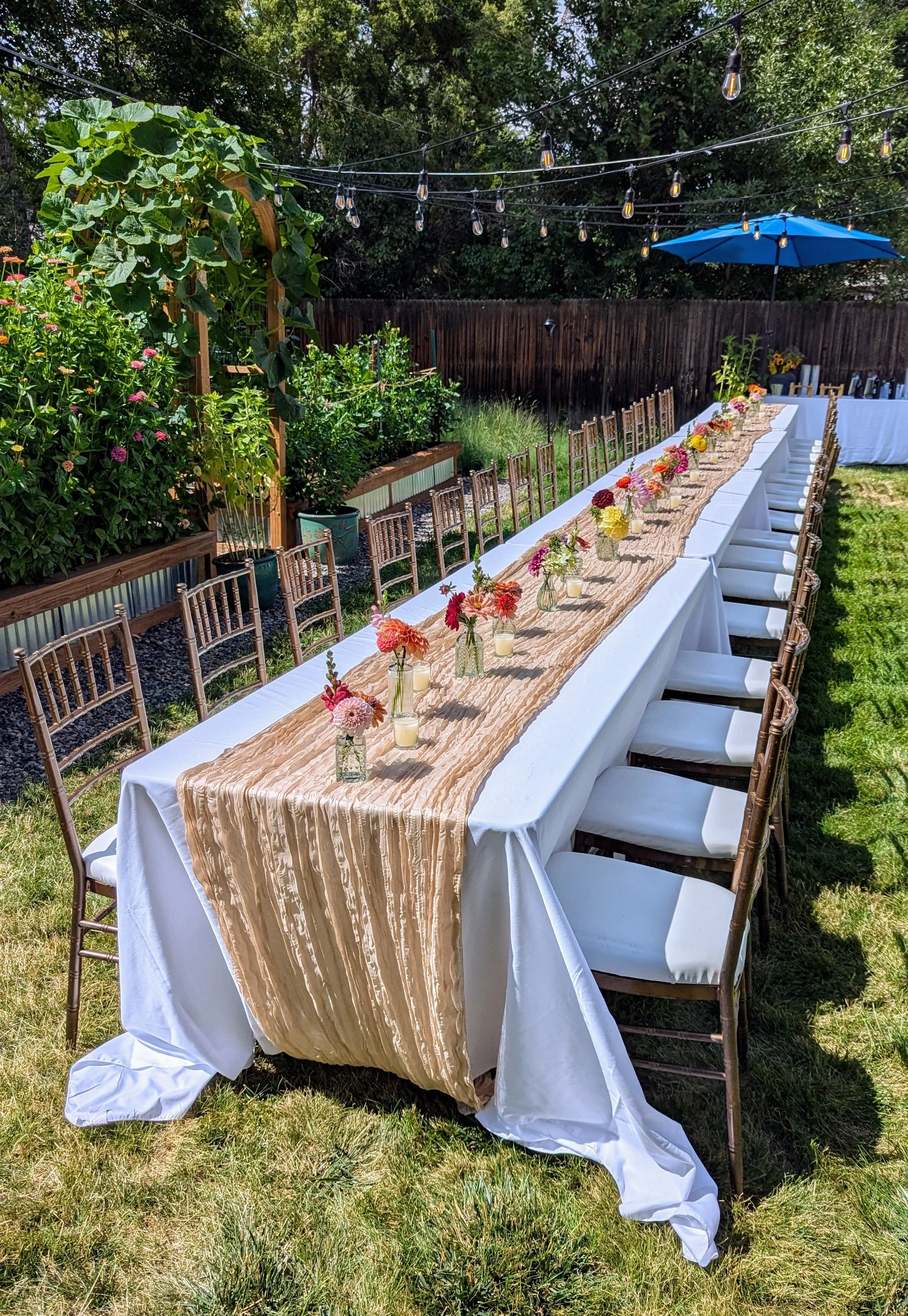 Long outdoor dining table decorated with flower arrangements and candles, set for an event in a backyard with string lights, a blue umbrella, and lush greenery.