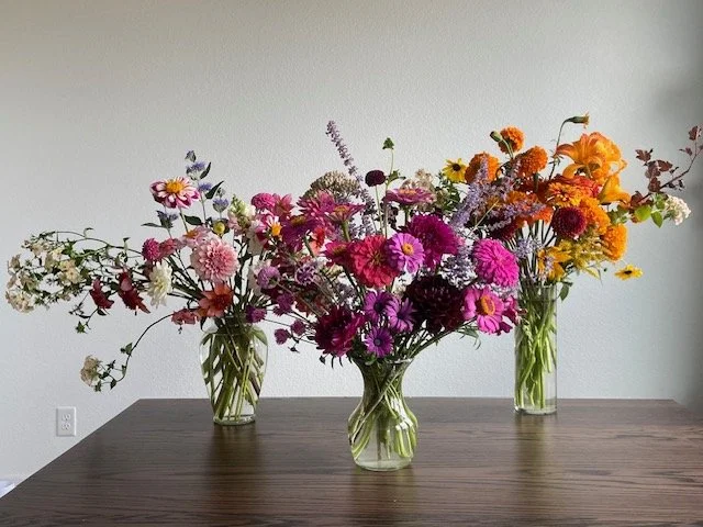 Three glass vases with colorful mixed flowers on a wooden table against a plain white wall.