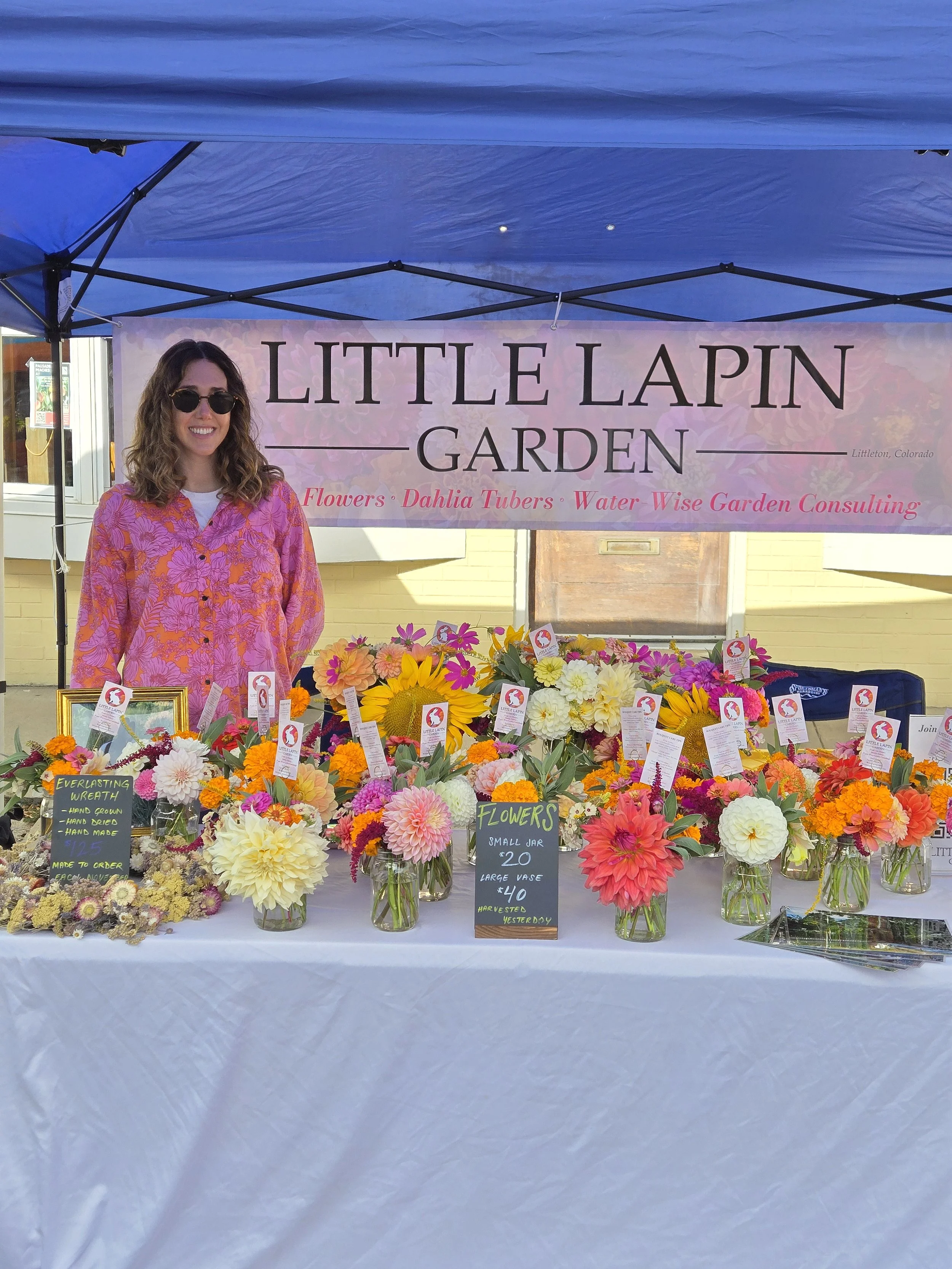 A woman standing behind a table full of colorful flowers at a flower shop booth named Little Lapin Garden, with a sign indicating flowers are sold in small jars and large vases, in Littleton, Colorado.