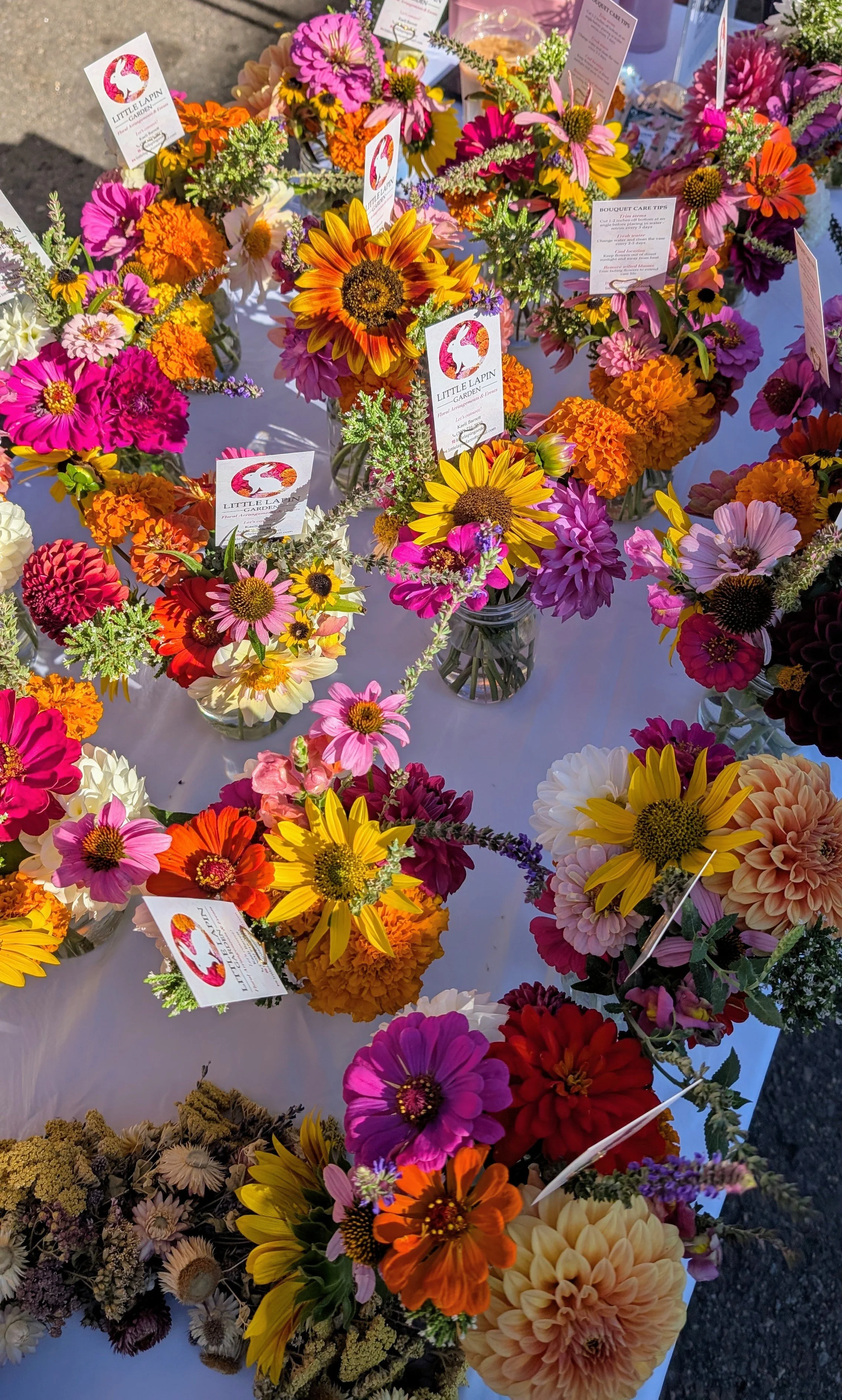 Colorful assortment of flower bouquets in glass jars, featuring various bright-colored flowers such as sunflowers, zinnias, and dahlias, displayed on a white table with small cards from Little Lapin Garden.