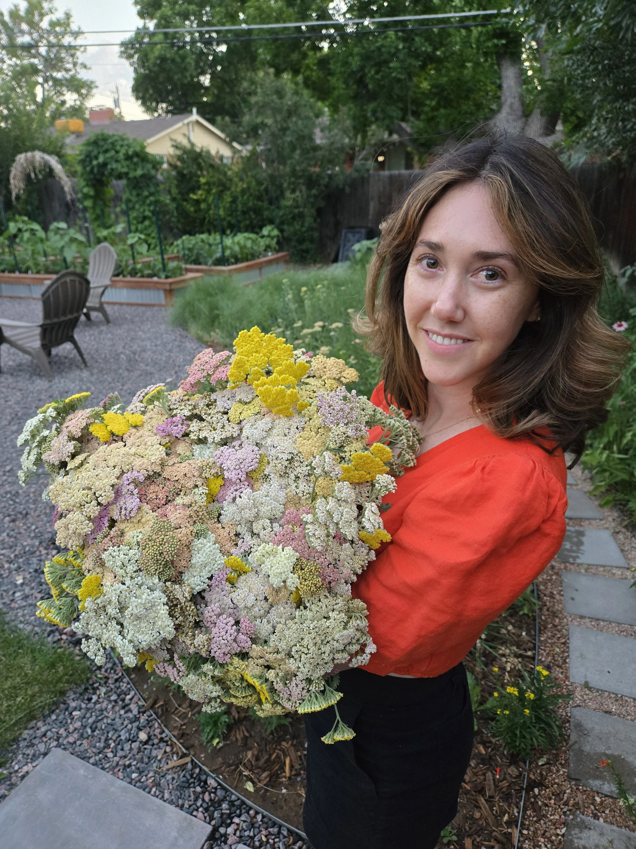 Woman with shoulder-length brown hair in a red blouse holding a large bouquet of colorful flowers in a backyard garden.