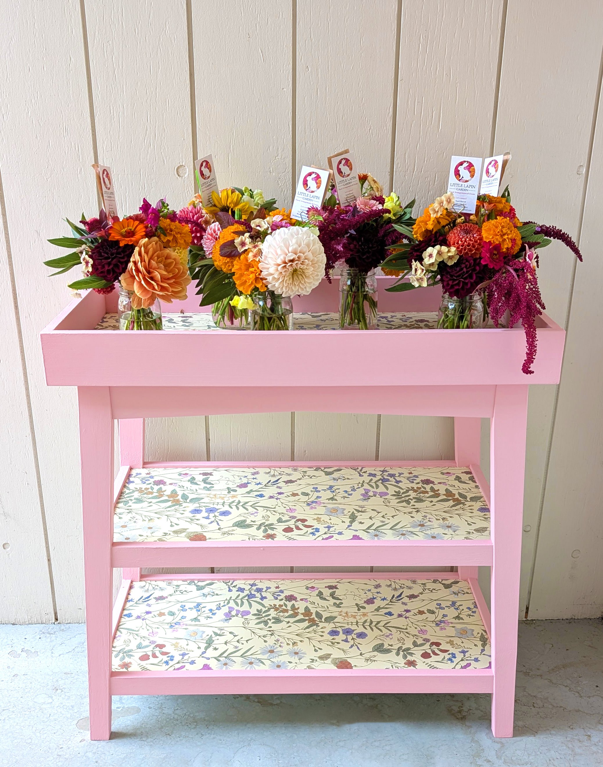 Pink wooden table with two shelves, decorated with floral wallpaper, holding five glass jars filled with colorful flower arrangements including dahlias, marigolds, and other blooms, against a light wooden wall.