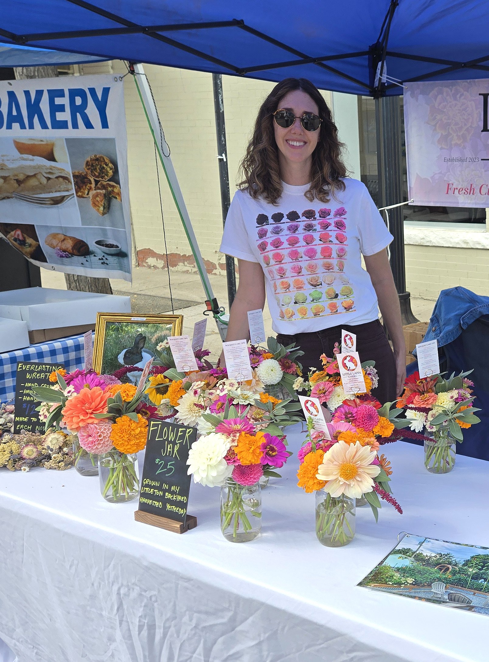 A woman with curly hair and sunglasses stands behind a table at an outdoor market. The table displays various colorful flower arrangements in glass jars with a small blackboard sign that reads 'Flower Jar'.