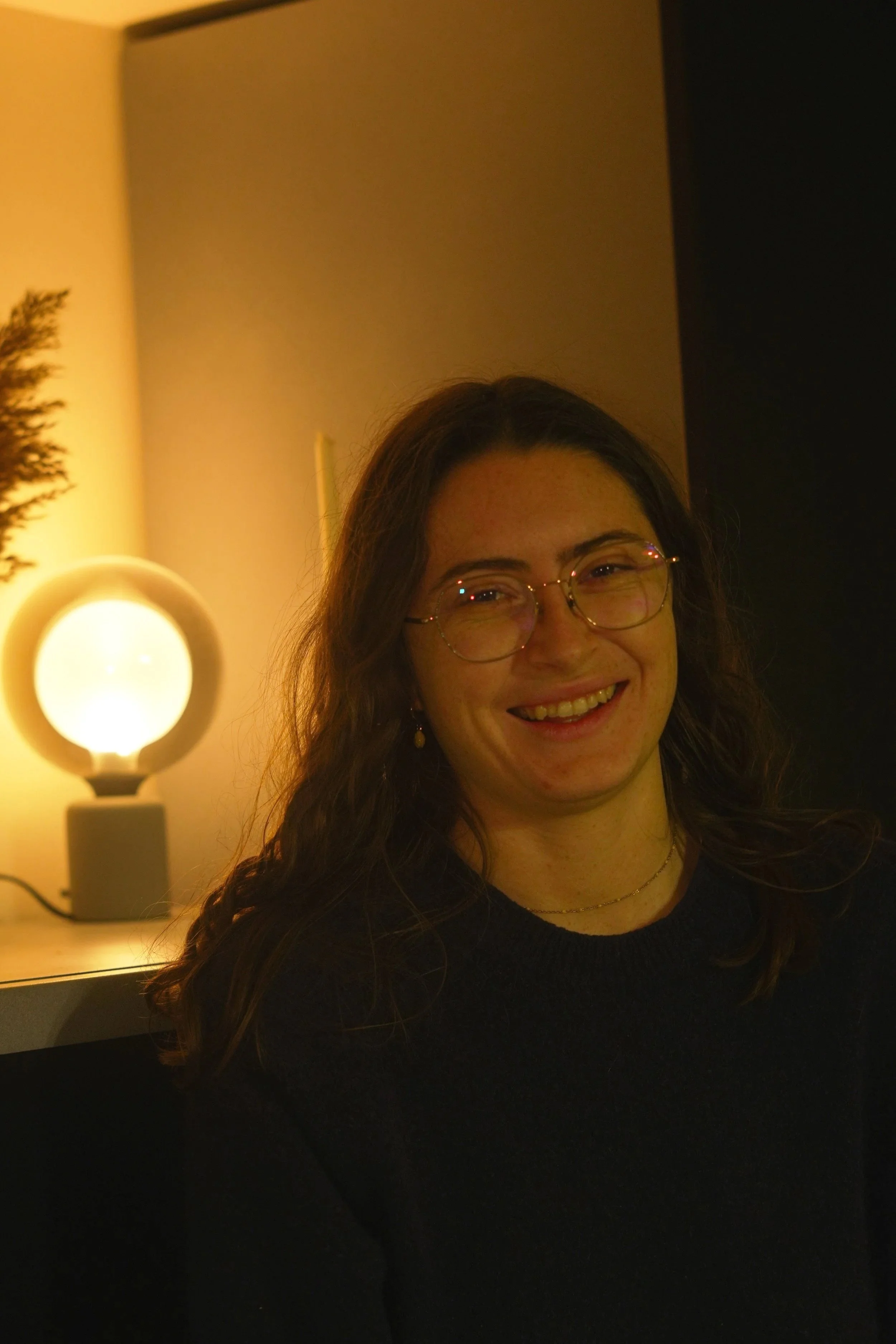 Une femme souriante portant des lunettes, assise près d'une lampe à lumière chaude dans une pièce sombre.