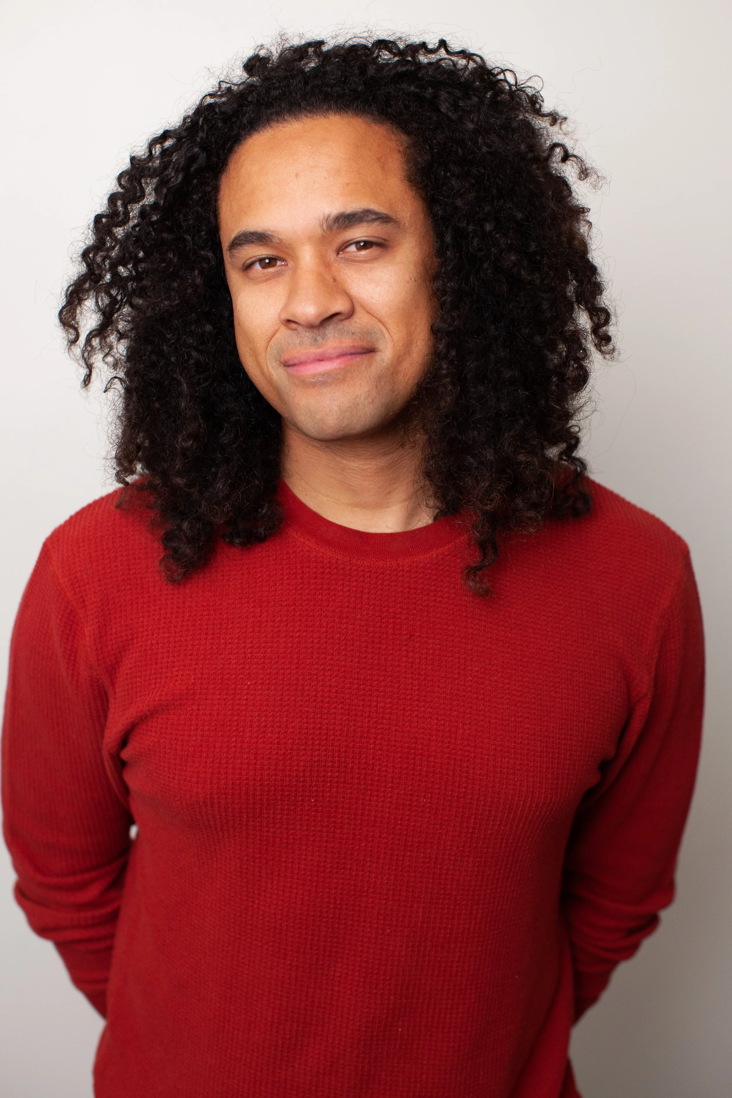 A man with long curly hair wearing a red sweater, standing against a plain white background.
