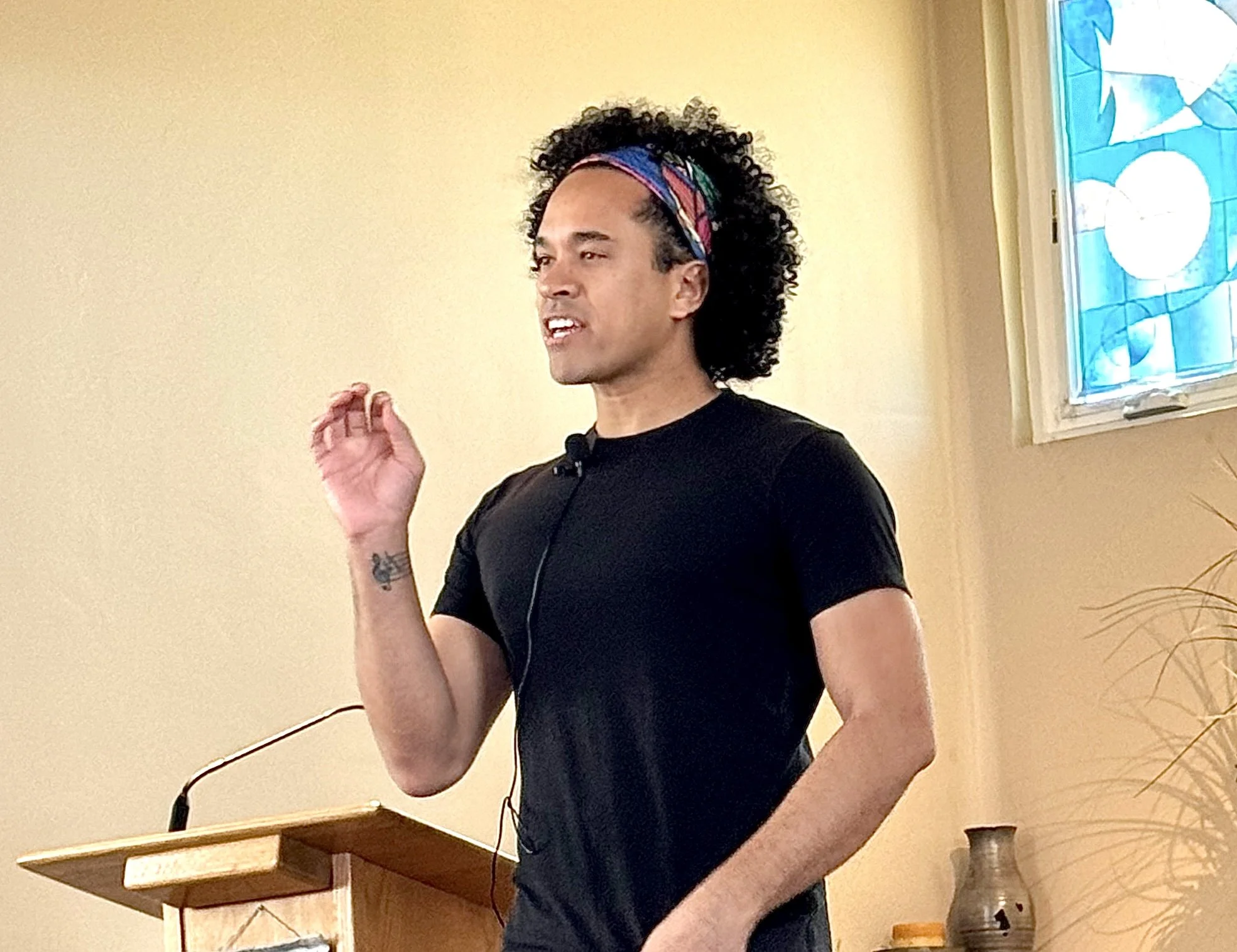A person with curly black hair, wearing a black t-shirt and a colorful headband, speaking at a podium indoors.