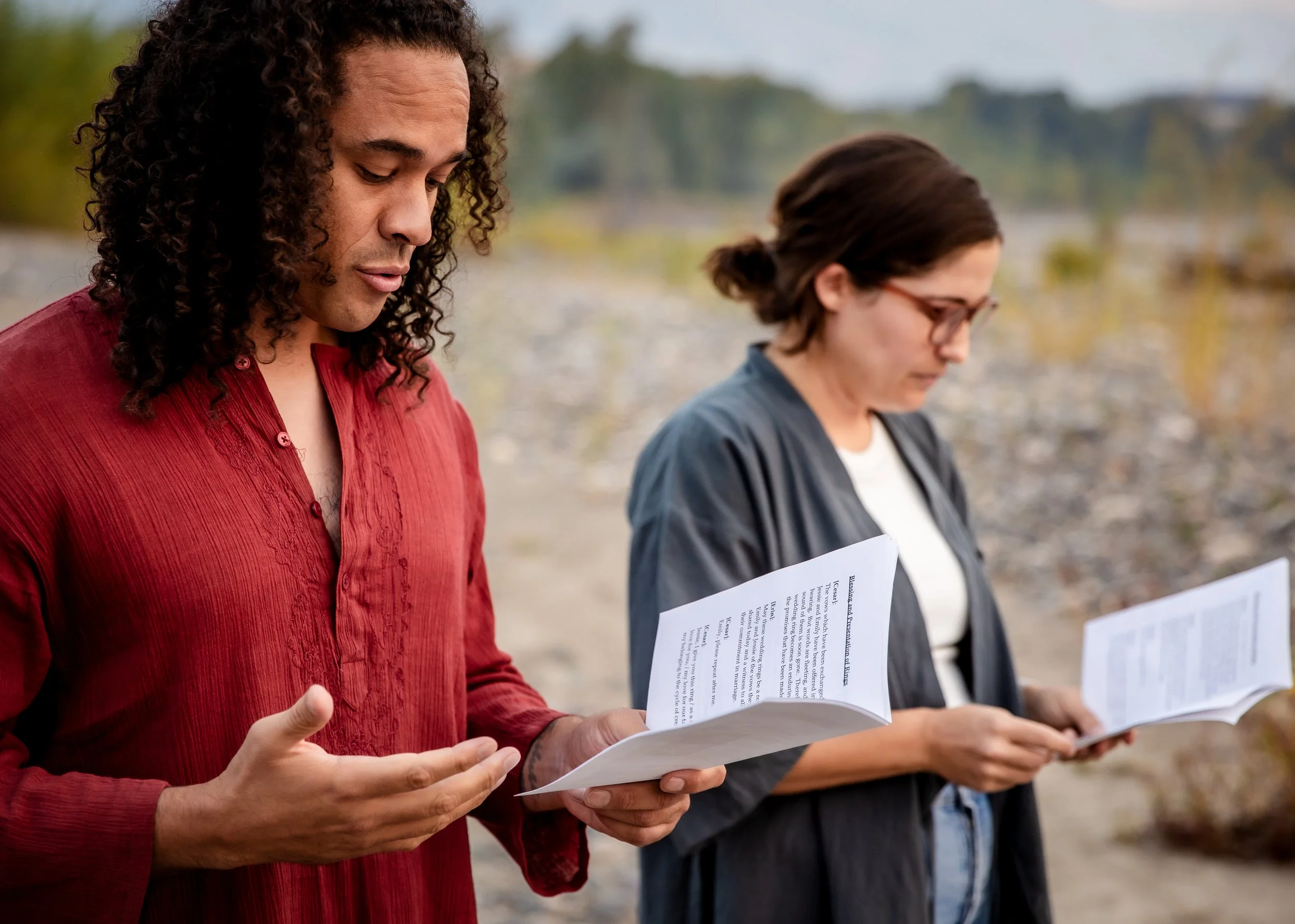Two women standing outdoors, reading papers, with a blurred natural background.