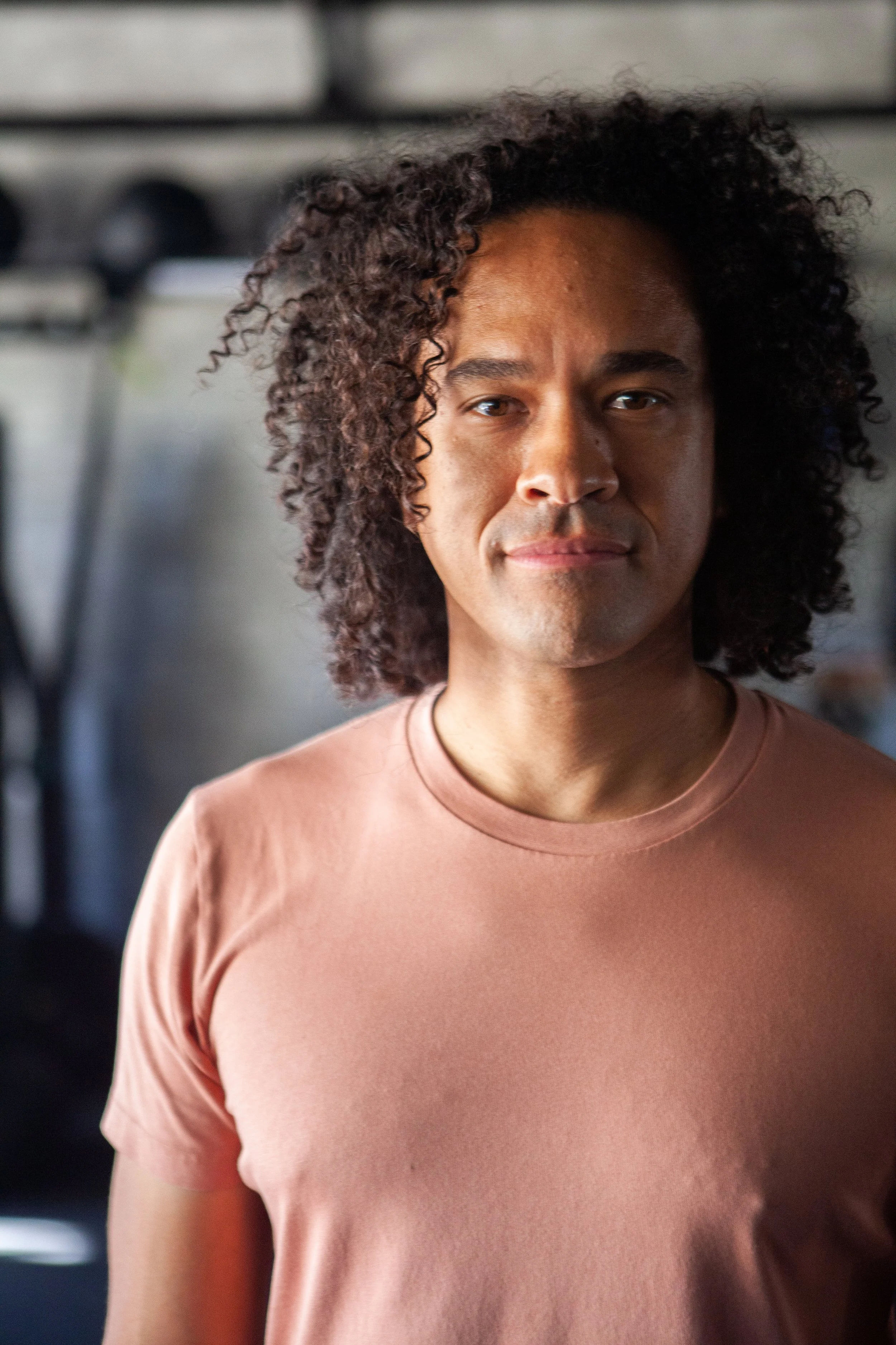 A man with curly hair wearing a pink T-shirt standing in a gym.