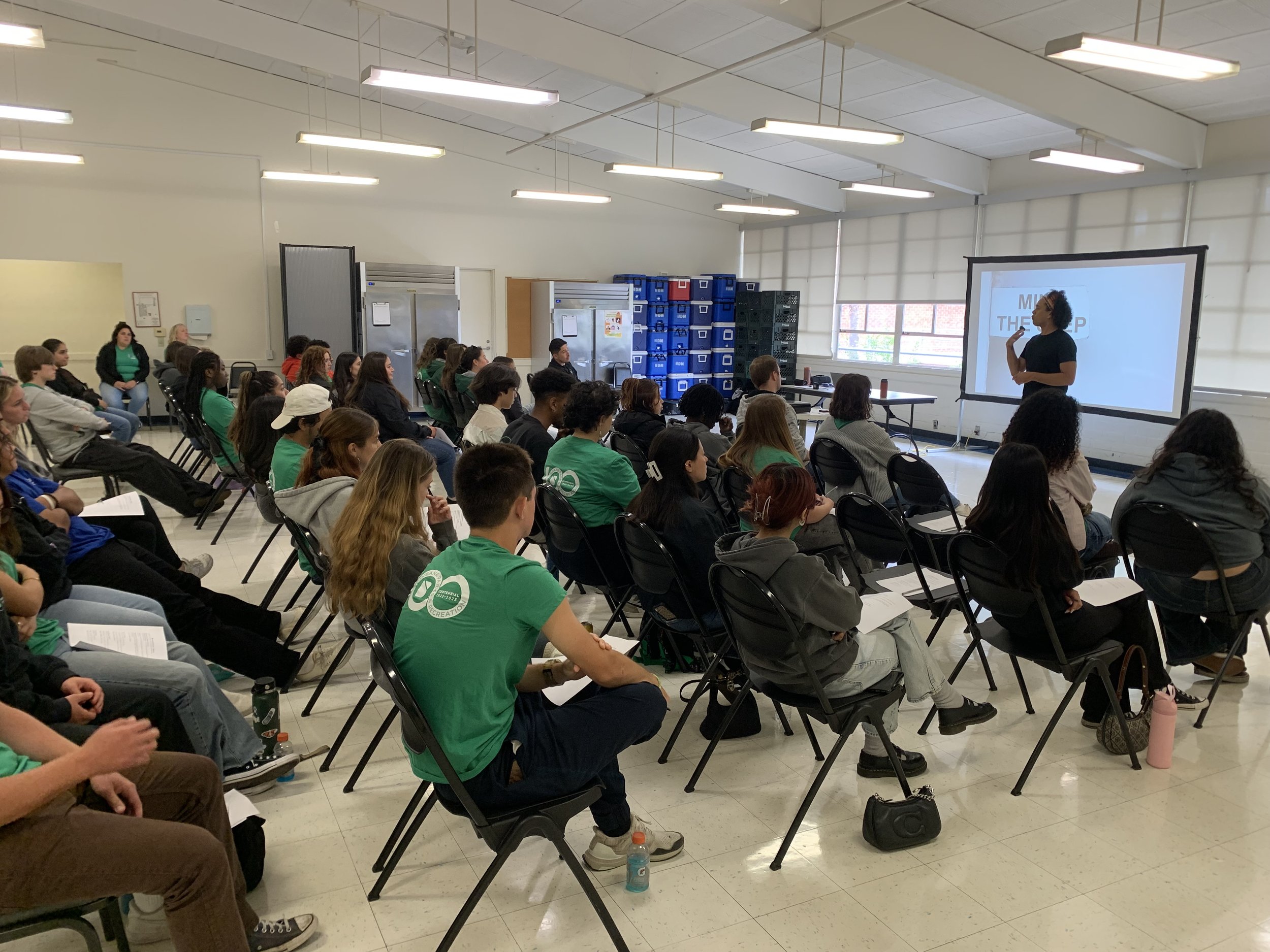 A diverse group of students seated in a classroom, watching a woman give a presentation at the front using a projector screen.