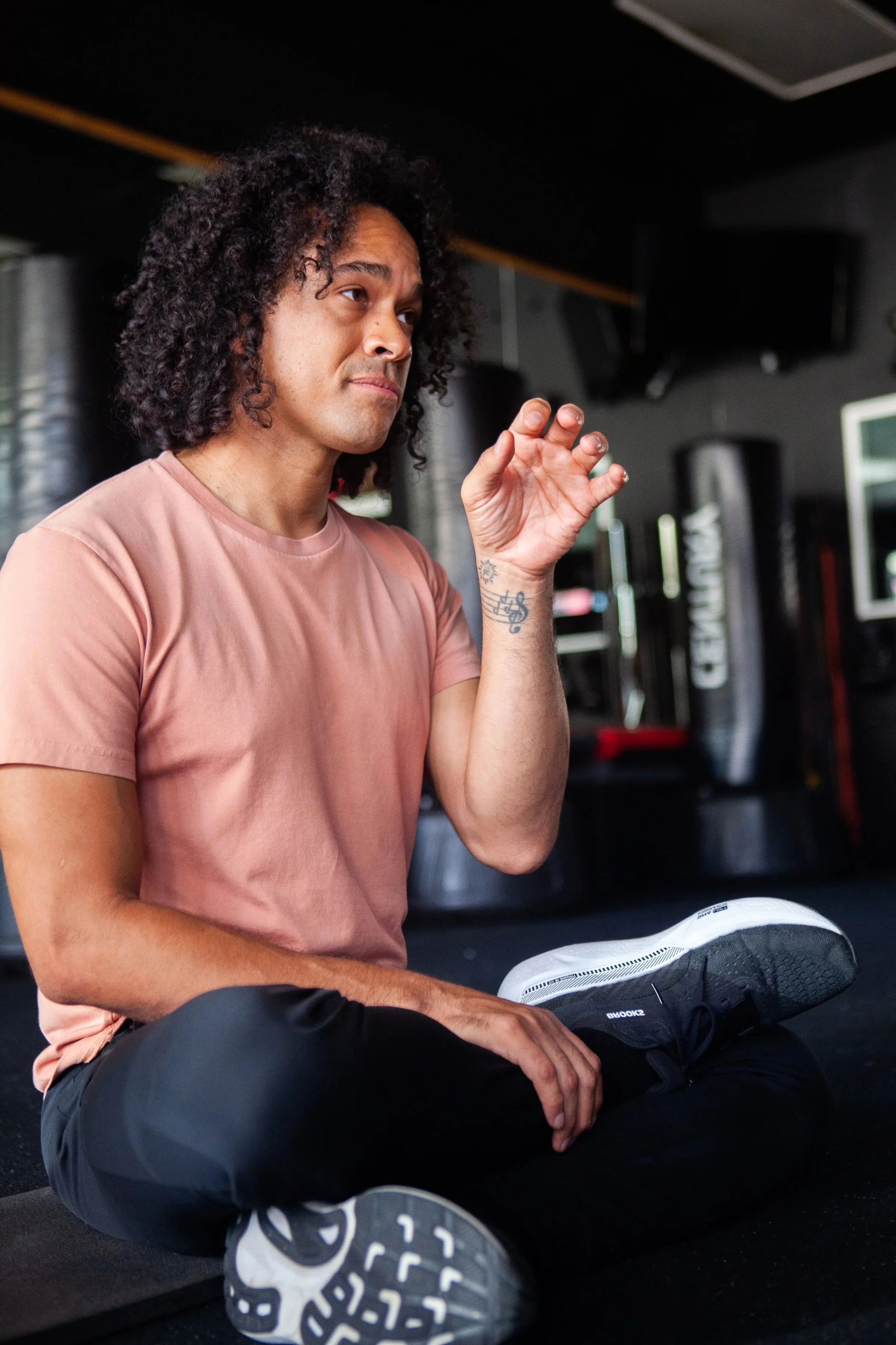 A man sitting cross-legged on the gym floor, with a sneaker resting on his knee, in a gym setting.
