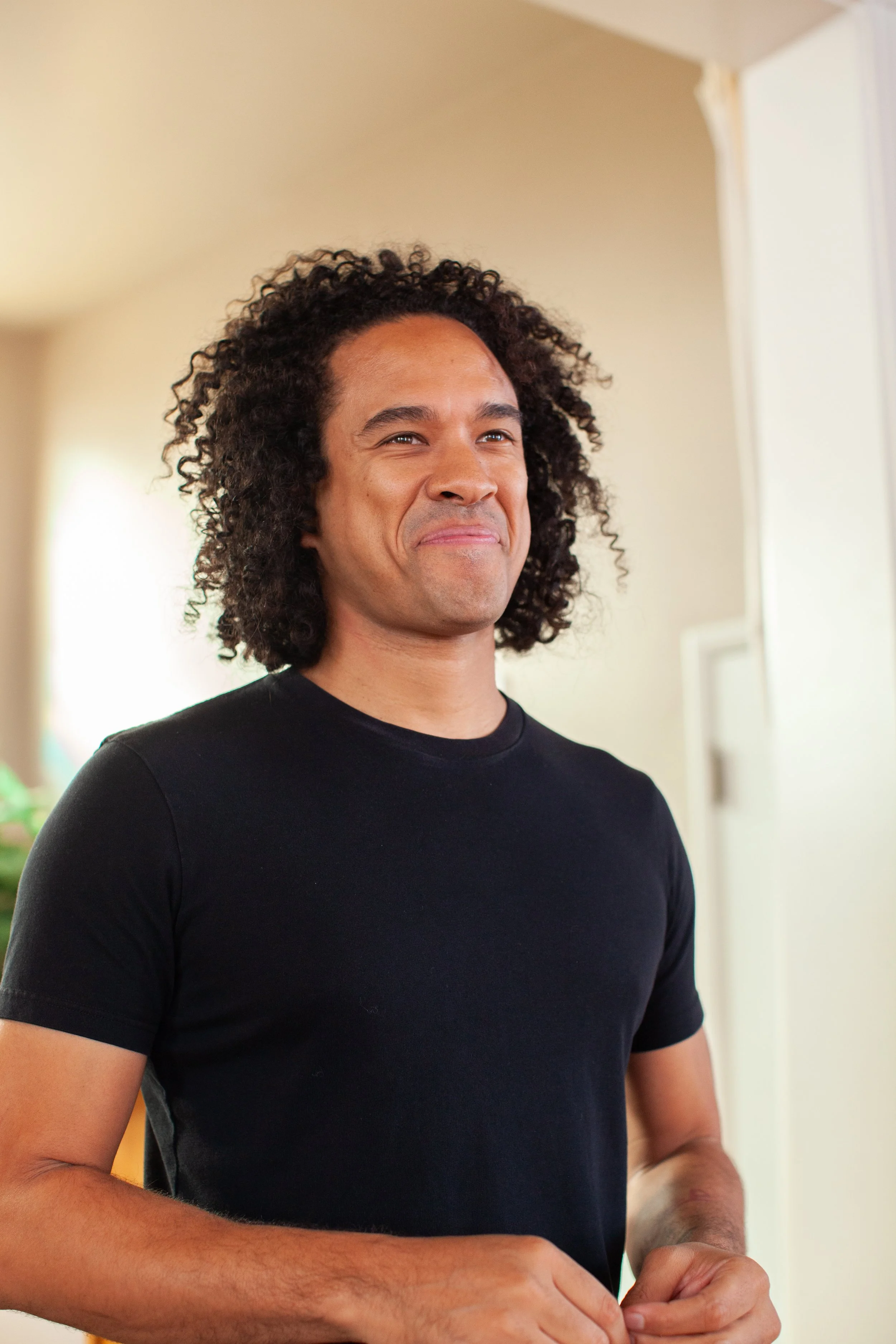 A young man with curly hair smiling while standing indoors, wearing a black t-shirt.