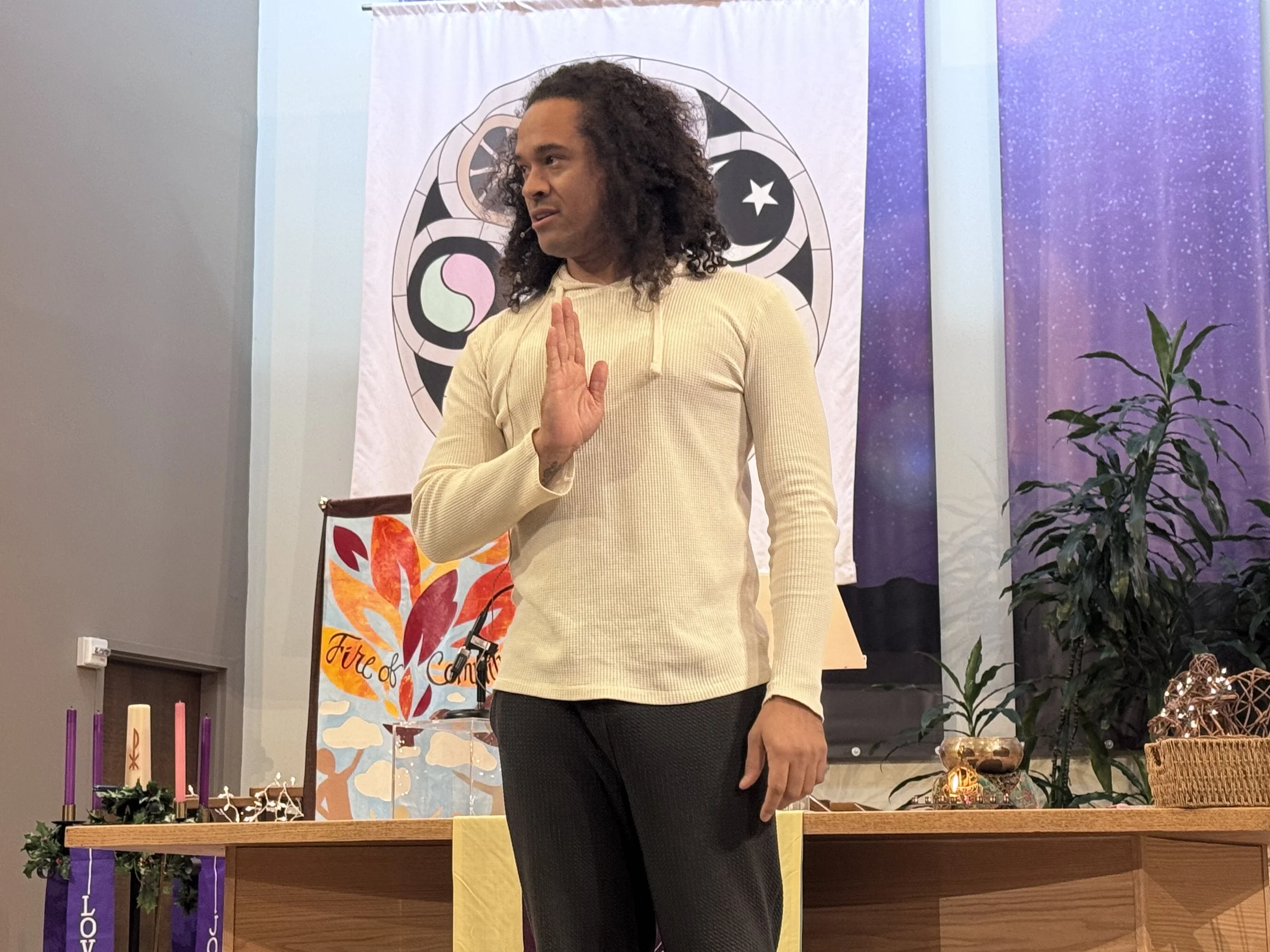 A man with curly hair wearing a beige hoodie gestures with his hand while standing in front of a colorful spiritual backdrop.